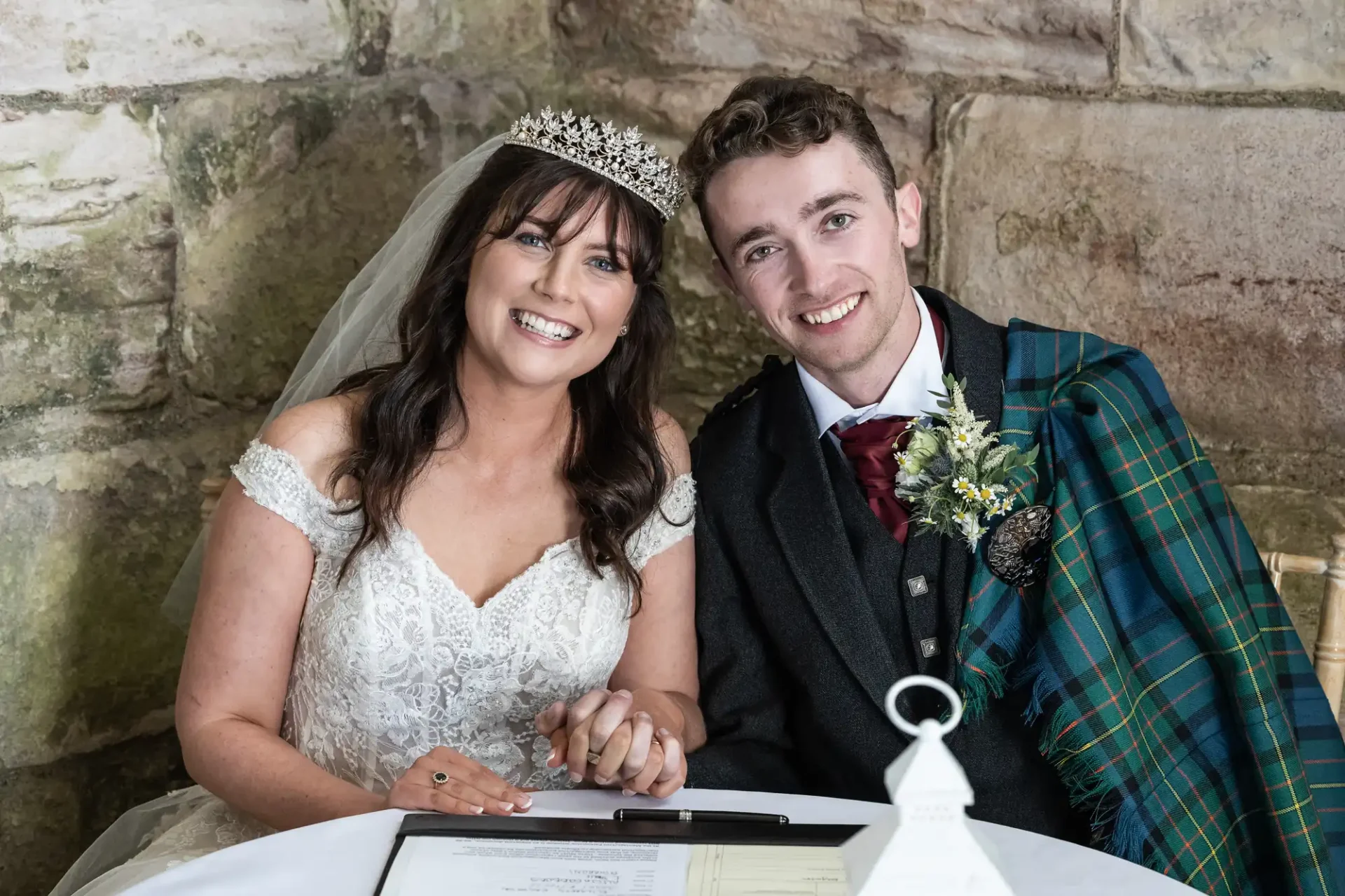 A bride and groom sit at a table, smiling. The bride wears a tiara and veil, and the groom wears a tartan shawl over his suit. They are holding hands with a document in front of them.