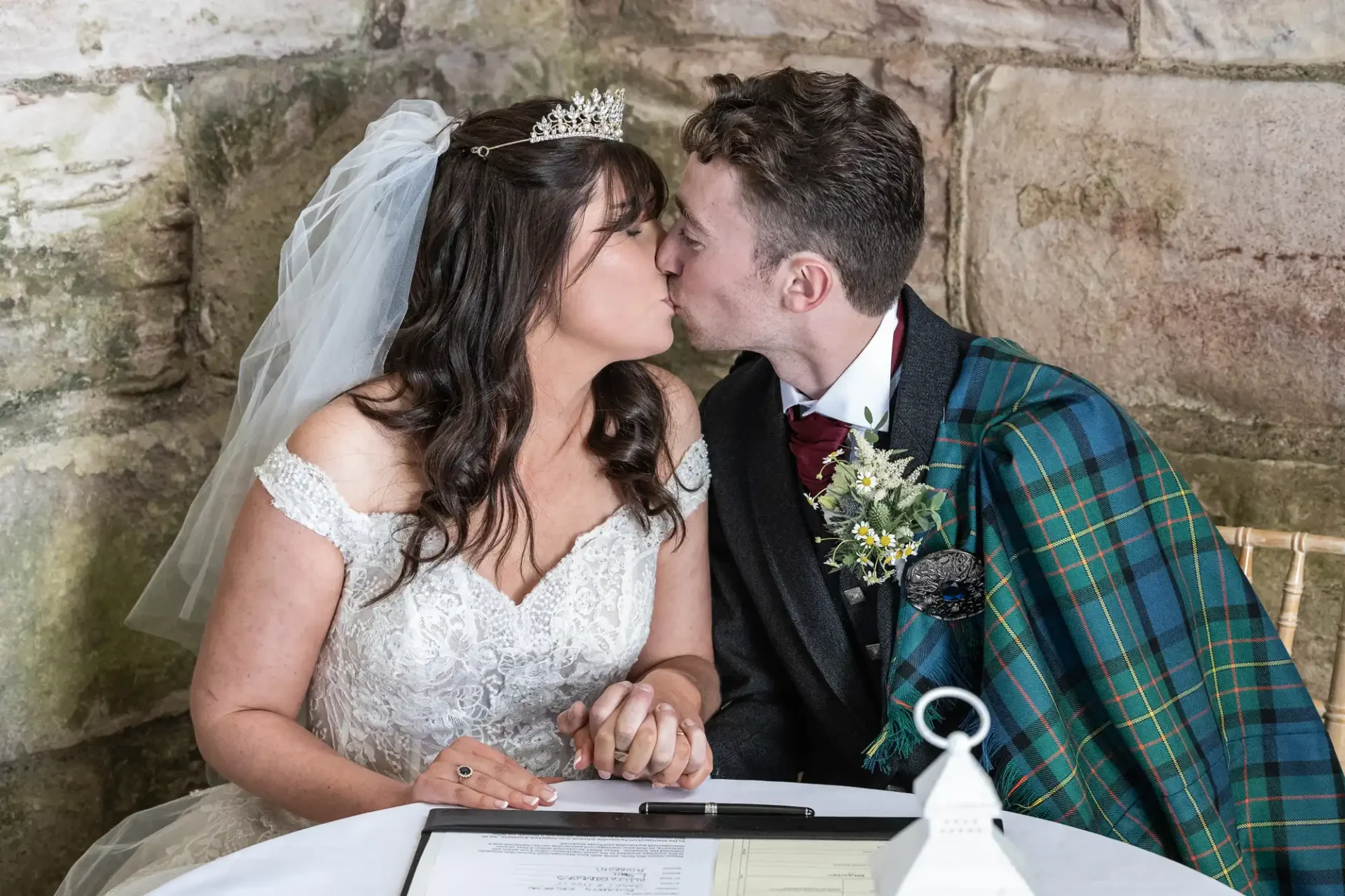 Bride and groom kiss while seated, holding hands. Bride wears a tiara and veil; groom dons a tartan sash. They are at a signing table against a stone wall.