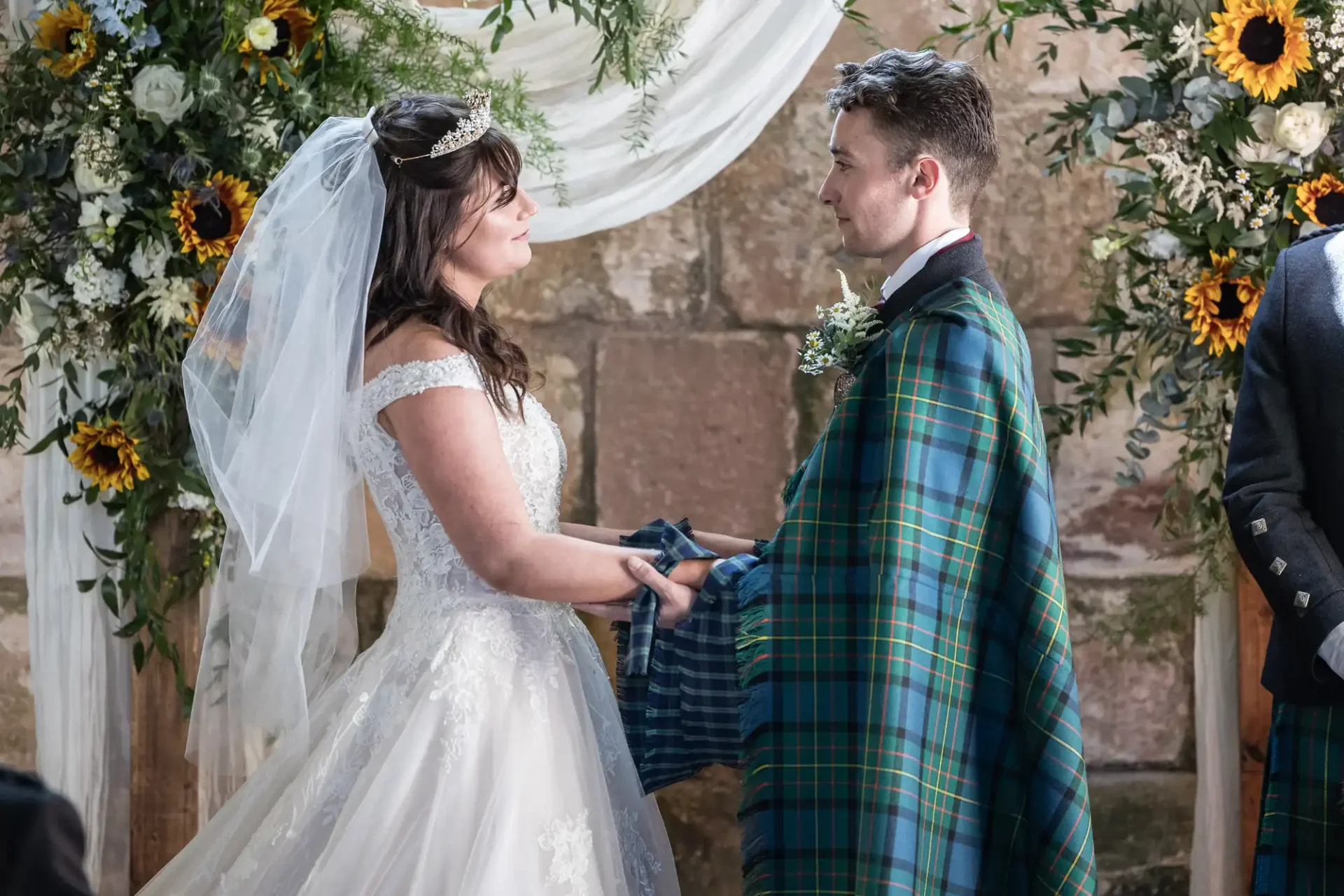 A bride and groom holding hands during a wedding ceremony. The bride wears a white dress and veil, while the groom wears a kilt with a tartan pattern. They stand before floral decorations.