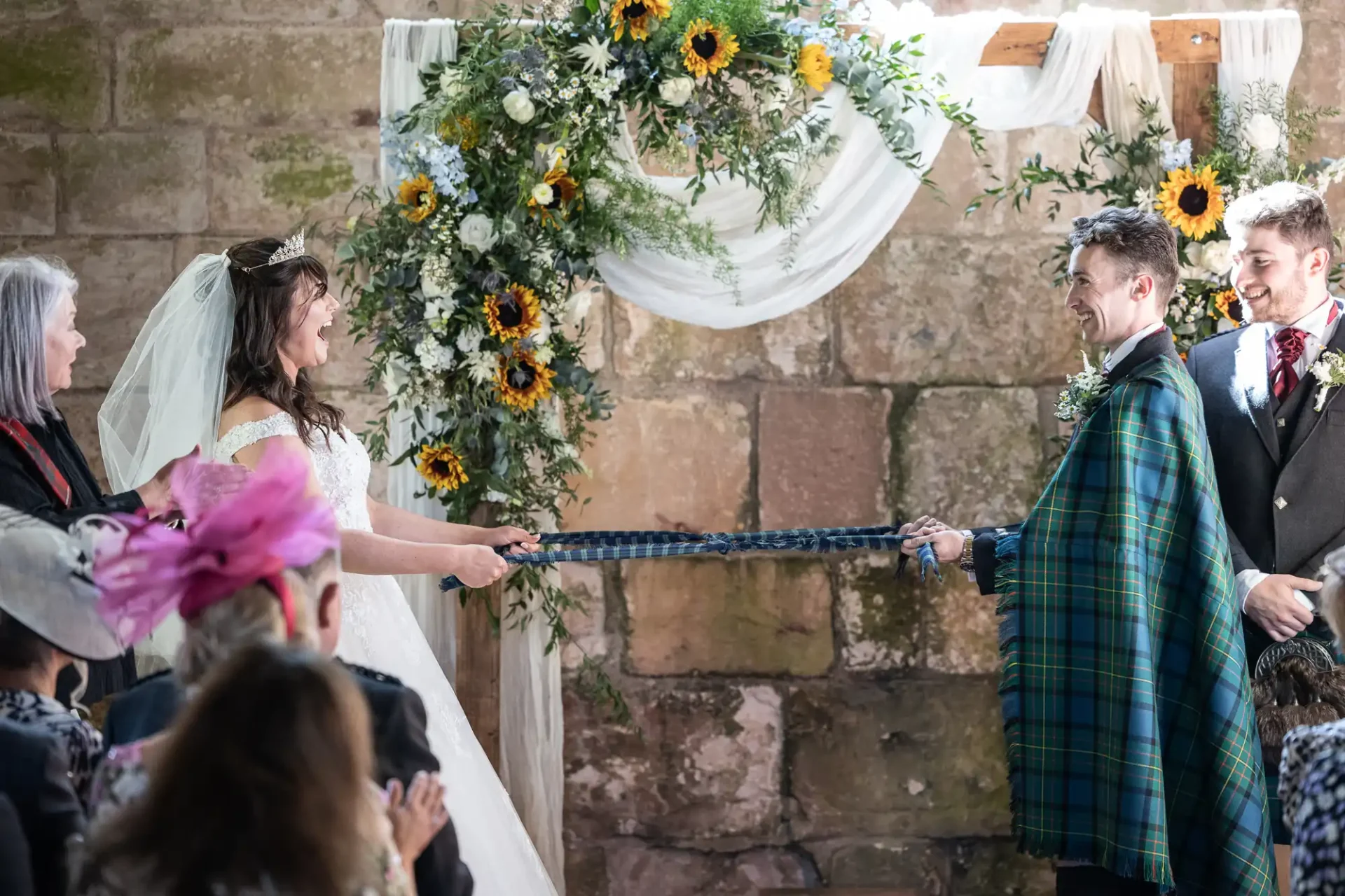 Bride and groom smiling during a handfasting ceremony, holding a blue rope, with floral decorations and guests in attendance.