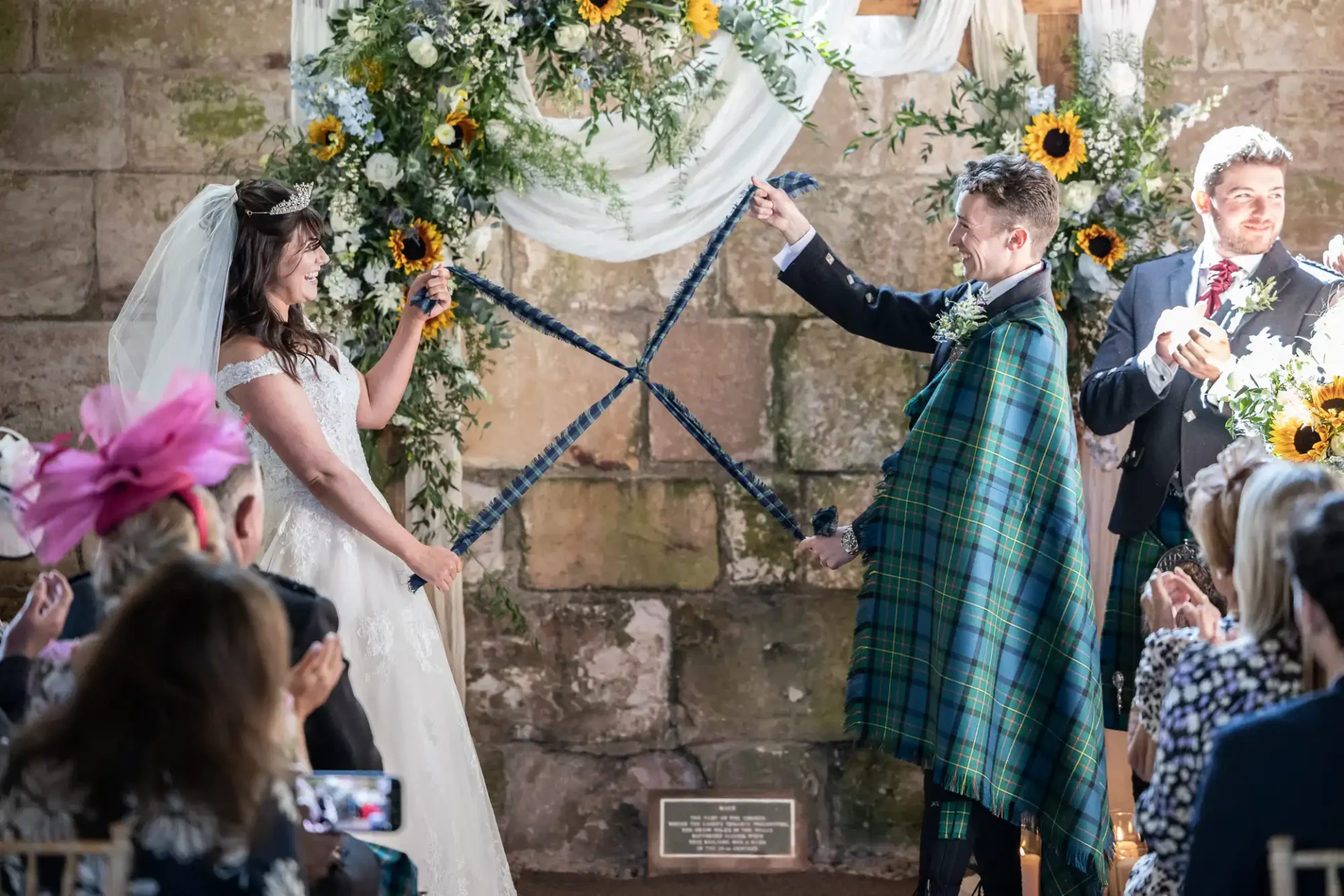 Bride and groom participate in a handfasting ceremony, holding tartan ribbons, under a floral arch with sunflowers and draped fabric. Guests watch in a stone-walled venue.