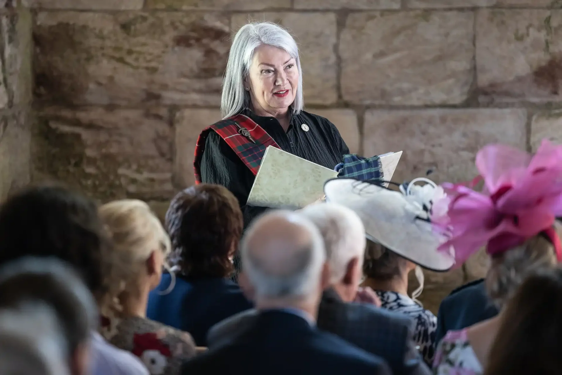 An older woman with white hair speaks to a seated audience. She is wearing a sash and holding papers, with a stone wall in the background. Some attendees wear hats with decorative details.