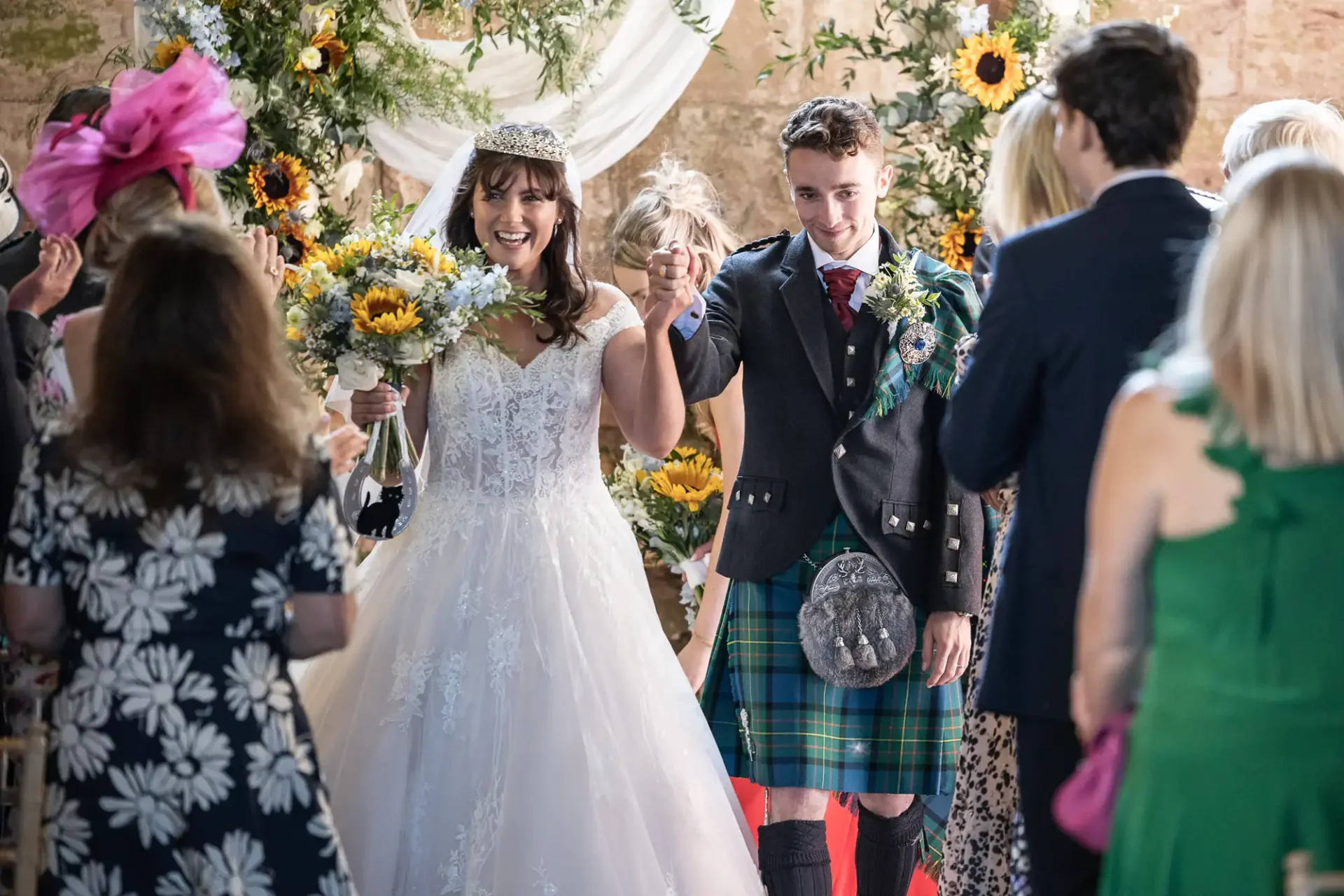 Bride and groom celebrate, holding hands amidst cheers. She wears a white gown with sunflowers; he wears a kilt. Guests in formal attire surround them in a decorated venue.