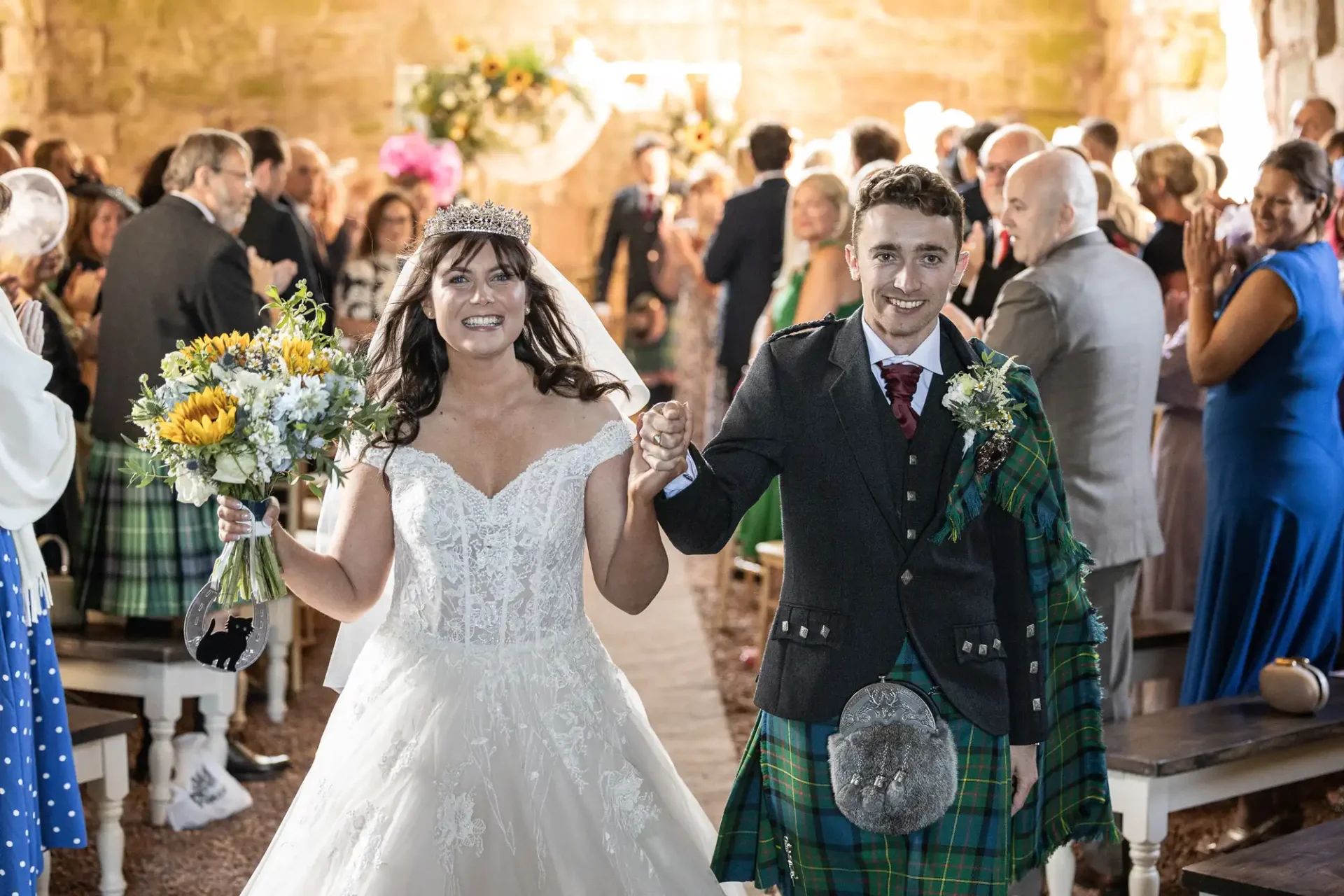 Bride and groom walking down the aisle, smiling, with guests clapping. Bride holds sunflower bouquet, and groom wears a green tartan sash.