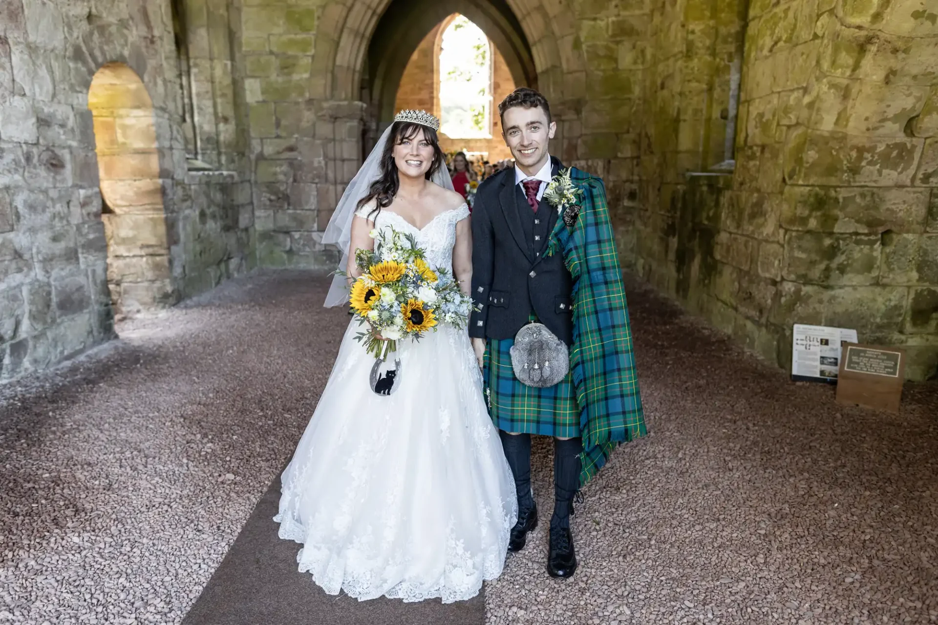 Bride in a white gown and groom in traditional Scottish attire stand together in a stone archway, with the bride holding a bouquet of sunflowers.