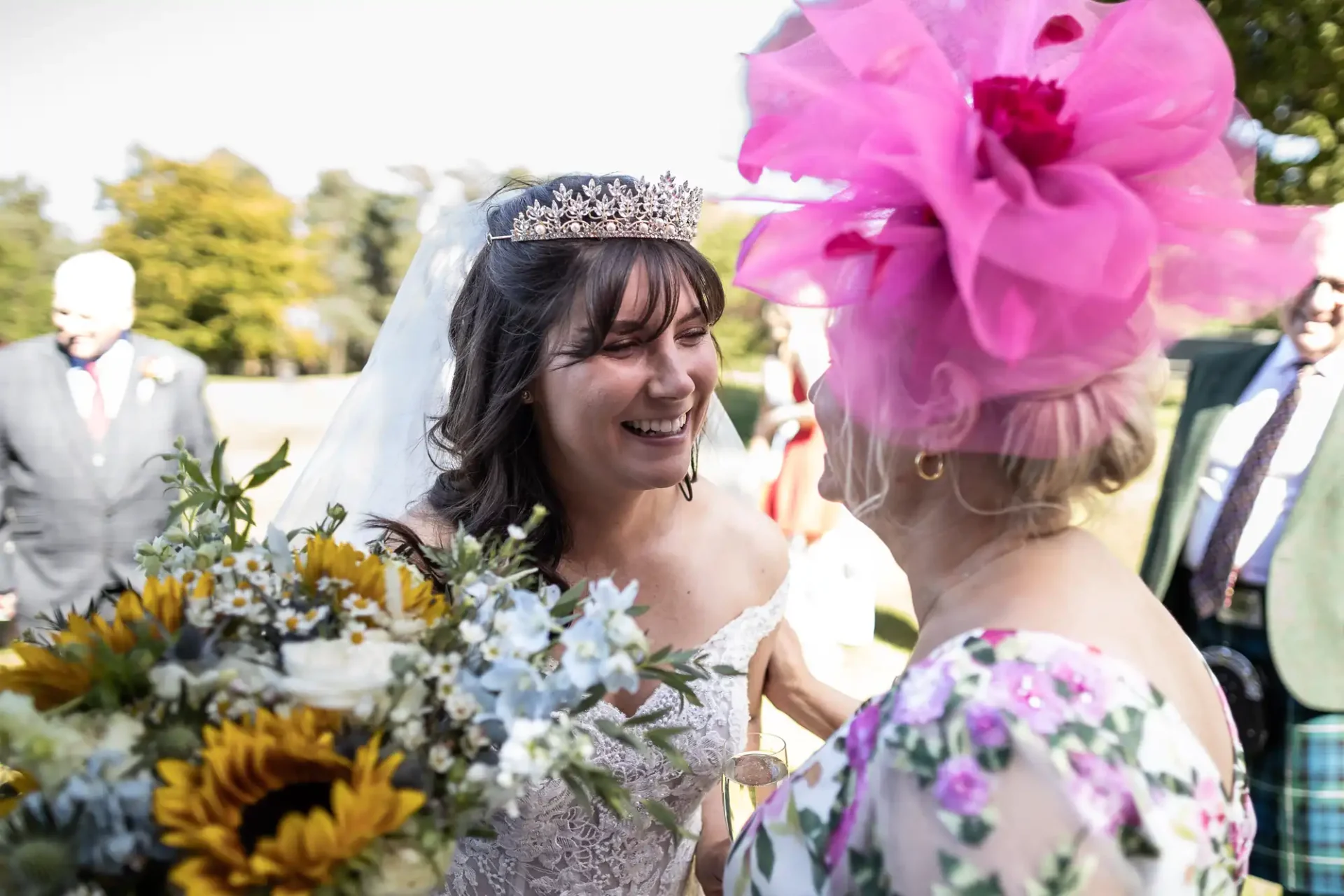 A bride in a white dress and tiara smiles while speaking with a woman in a pink hat and floral dress, holding a bouquet of sunflowers and other flowers outdoors.