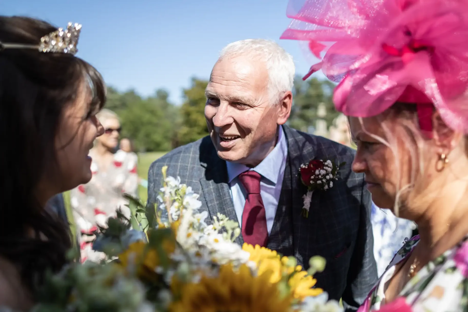 An older man in a suit with a boutonniere stands outdoors, talking to two women dressed in formal attire. One woman wears a large pink hat. Sunflowers and greenery are in the foreground.