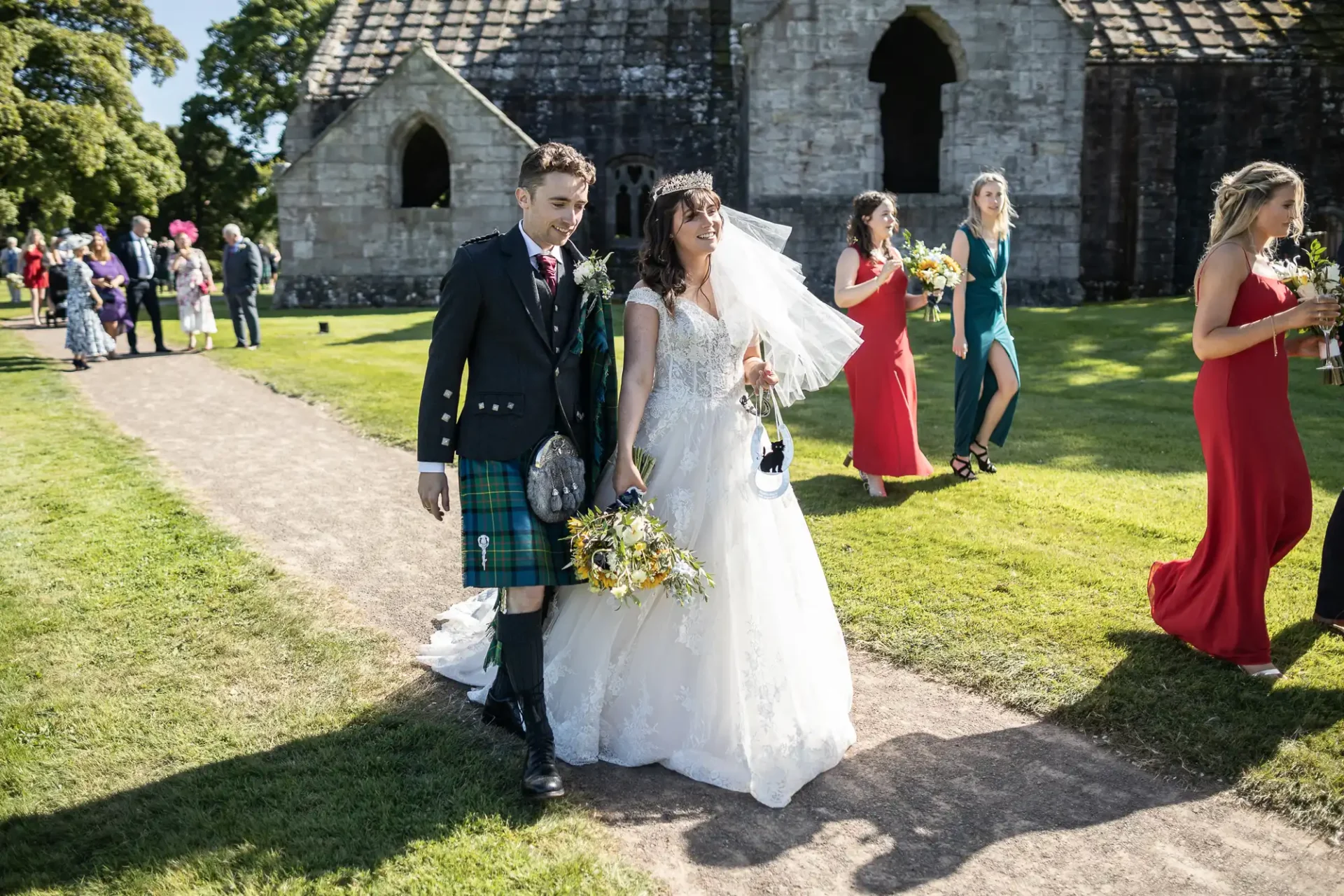 A bride and groom walk down a path at an outdoor wedding. The groom wears a kilt and the bride holds a bouquet. Guests are seen in the background near an old stone building.