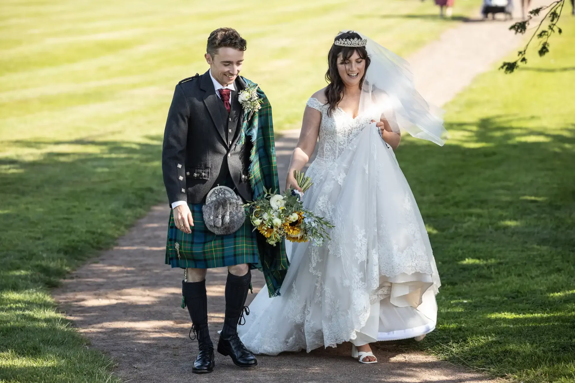 A couple walks on a grassy path, with the man in a kilt and the woman in a white wedding dress holding a bouquet.