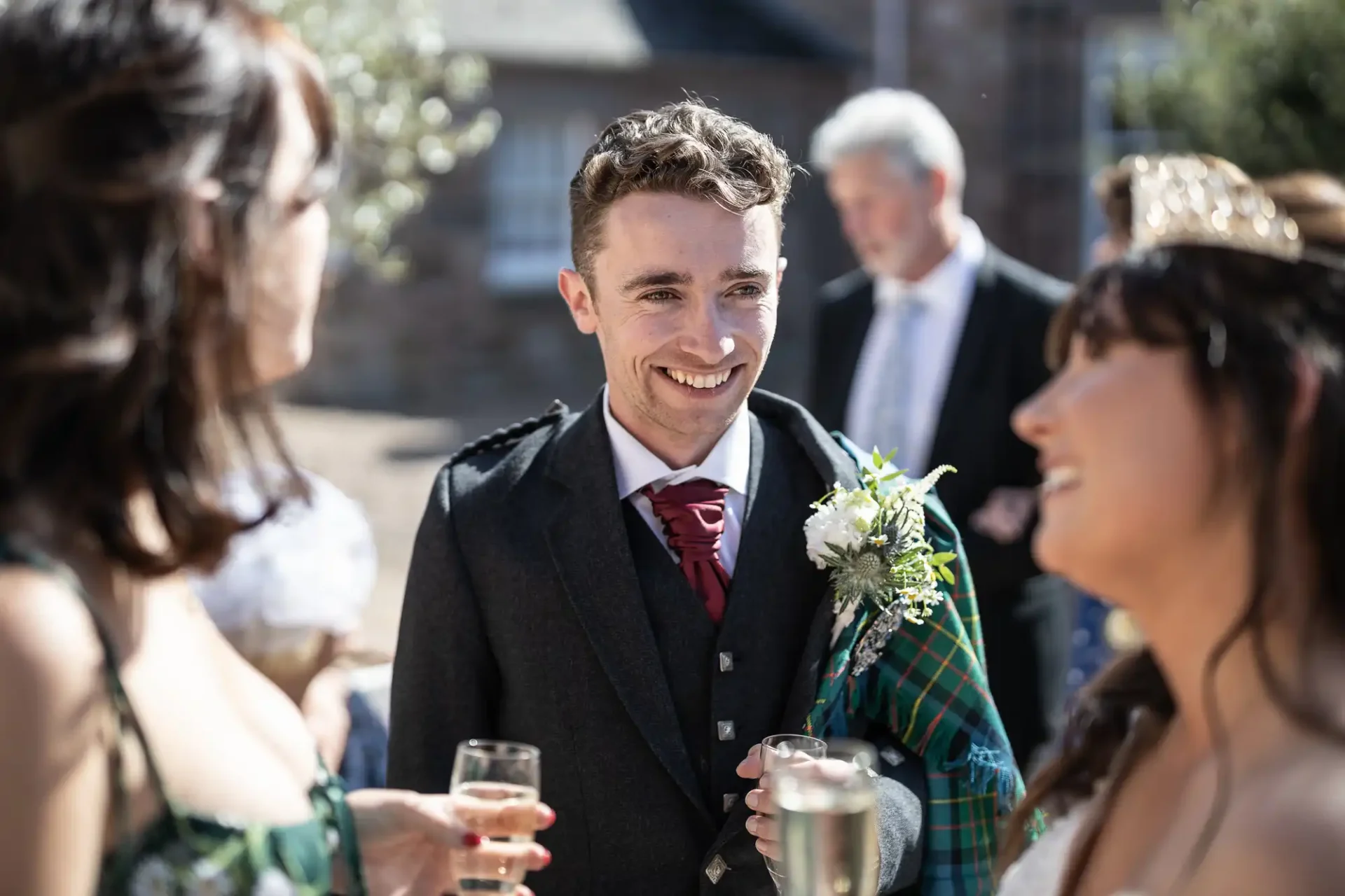 A man in formal attire, holding a plaid cloth and a drink, smiles at two women during an outdoor gathering.