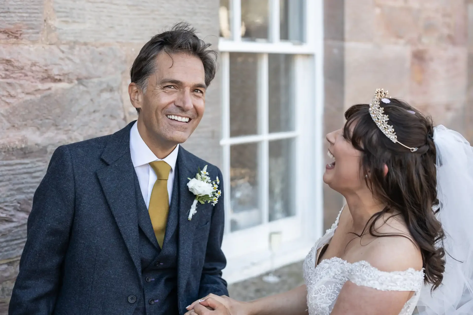 A man in a suit smiles while holding hands with a woman in a wedding dress. They are standing in front of a stone wall with a window.