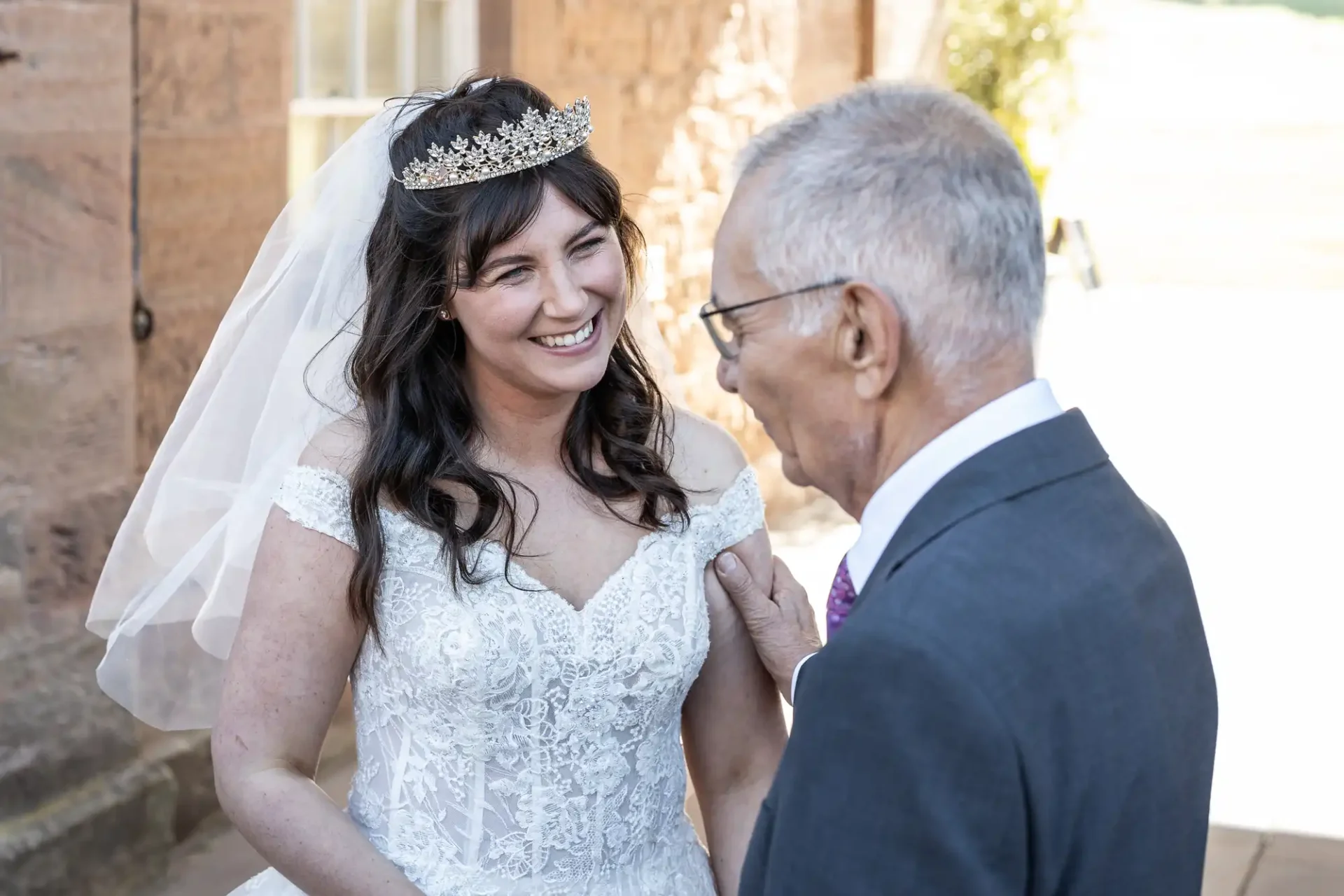 Bride in a white dress and tiara smiles at an elderly man in a suit outdoors.