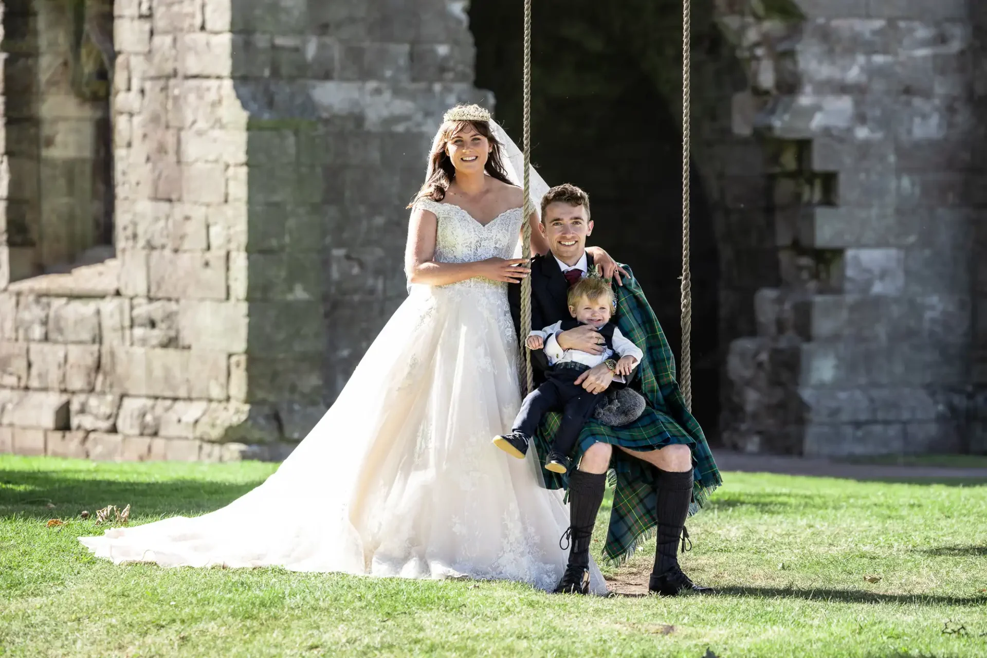 A bride in a white gown and veil stands next to a groom in a kilt sitting on a swing with a young boy. They are outdoors in front of a stone ruin.