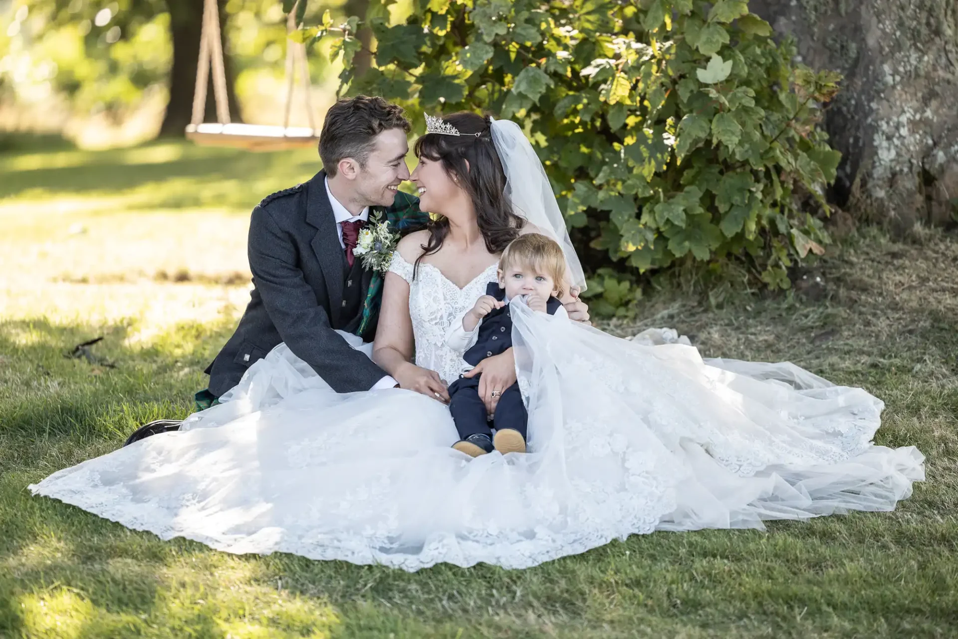 Bride and groom sitting on grass with a toddler in a wedding dress. They are posing close together in a sunlit outdoor setting.