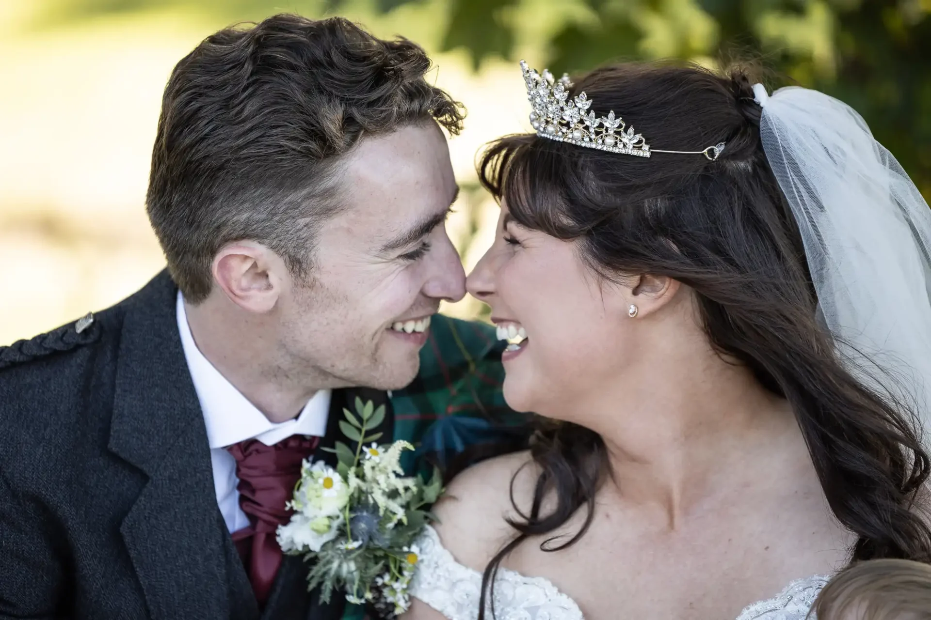 A bride and groom are sitting close, smiling at each other. The bride wears a tiara and veil, and the groom is in a suit with a floral boutonniere.