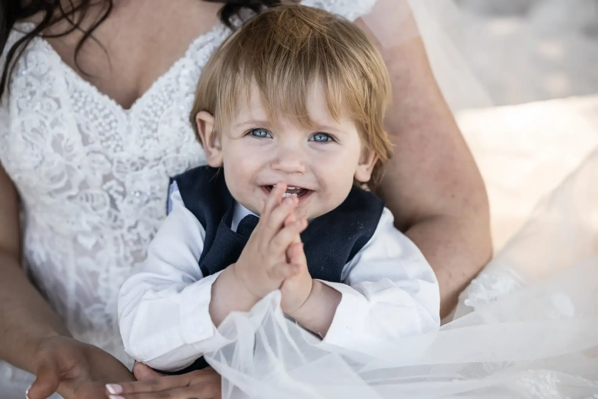 A smiling toddler wearing a white shirt and navy vest clasps hands together while sitting on a woman's lap in a white dress.