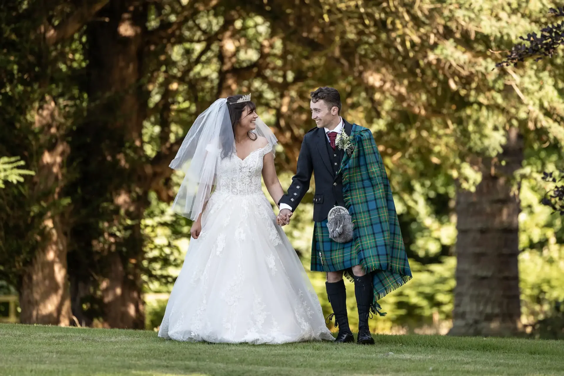 A bride in a white gown and a groom in a tartan kilt walk hand in hand on grass, surrounded by trees.