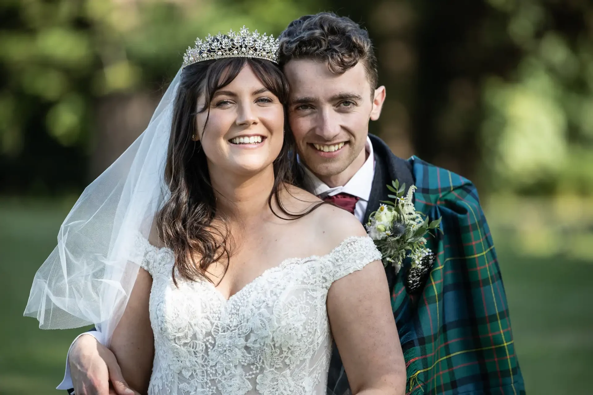 Bride and groom smiling together outdoors; she wears a lace dress and tiara, he wears a suit with a plaid sash.