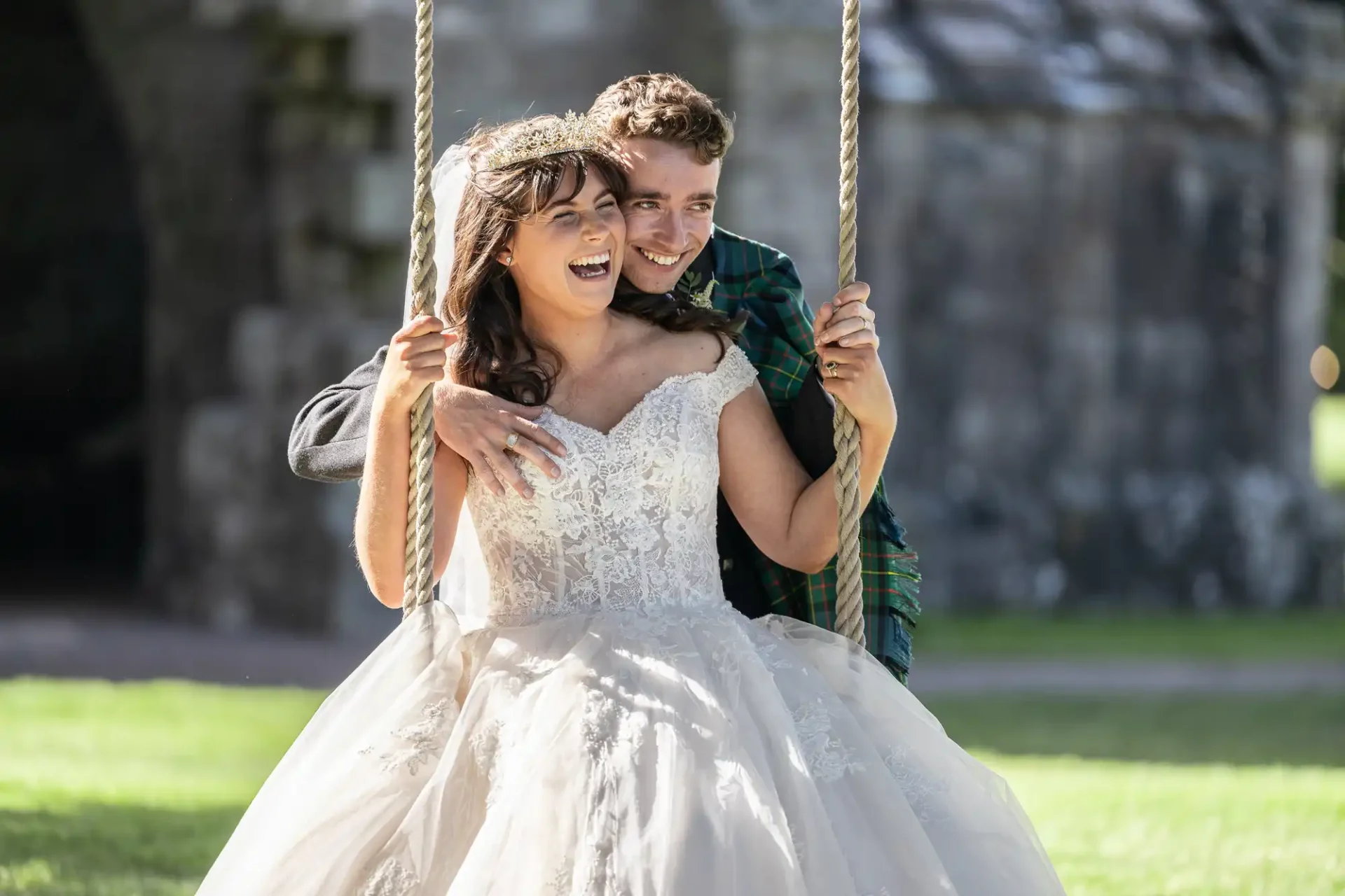 A bride in a white lace gown and a groom in a plaid scarf smile while standing on a swing.