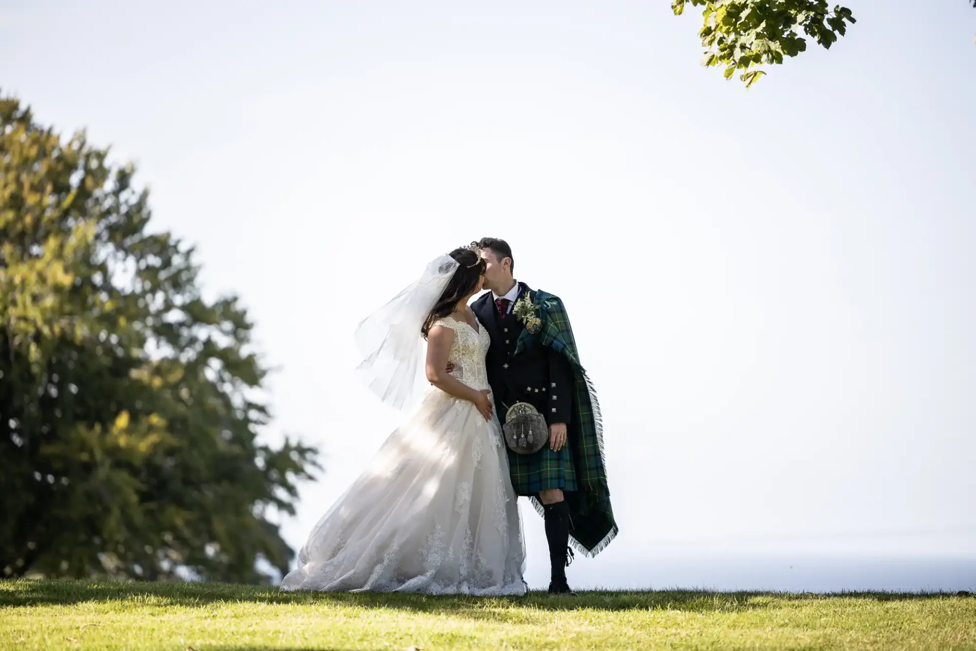 Bride and groom kissing under a tree, with the groom wearing a kilt and the bride in a wedding dress.