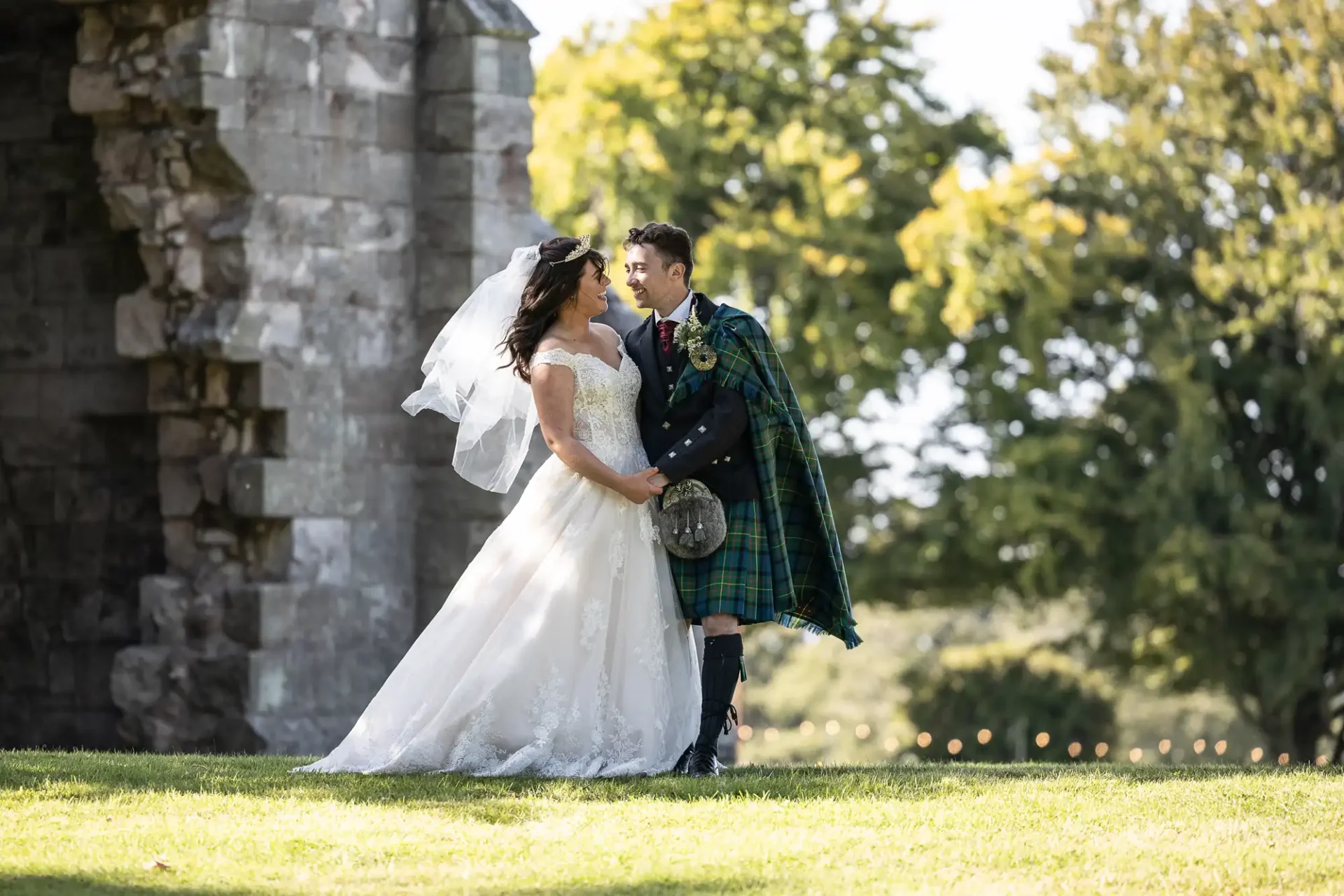 Bride and groom standing outdoors, holding hands and smiling at each other. Groom wears a kilt and tartan attire; bride wears a white gown. Stone structure and trees in the background.