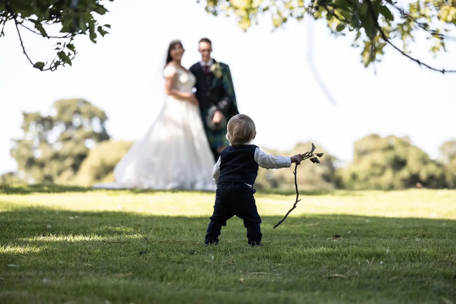 Child holding a flower and stick on grass, with a distant couple in wedding attire out of focus in the background.