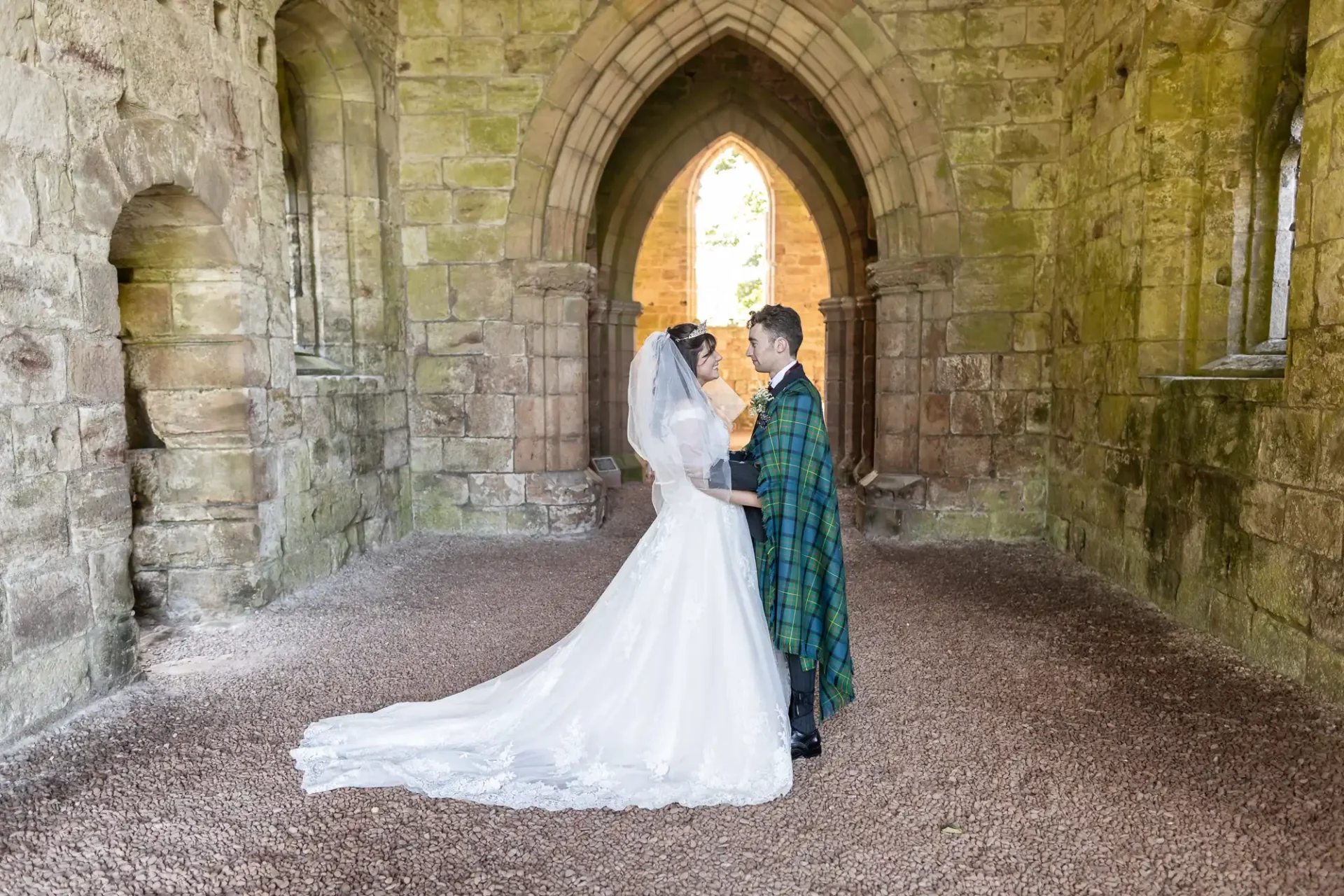 Bride in a white gown and groom in a green tartan attire stand facing each other inside a stone archway of an old building.