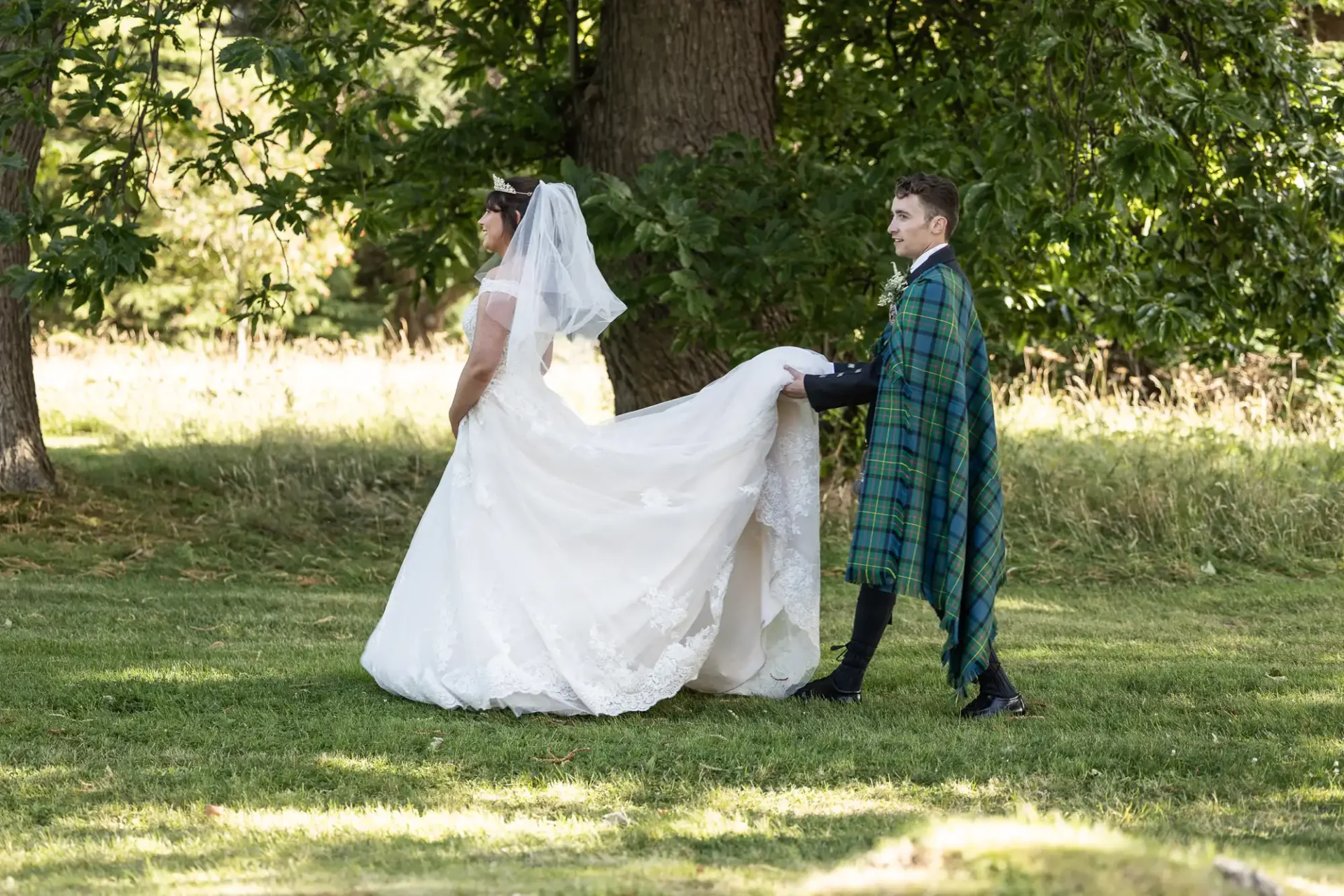 A groom in a green tartan outfit holds the train of a bride's white dress as they walk on grass in a wooded area.