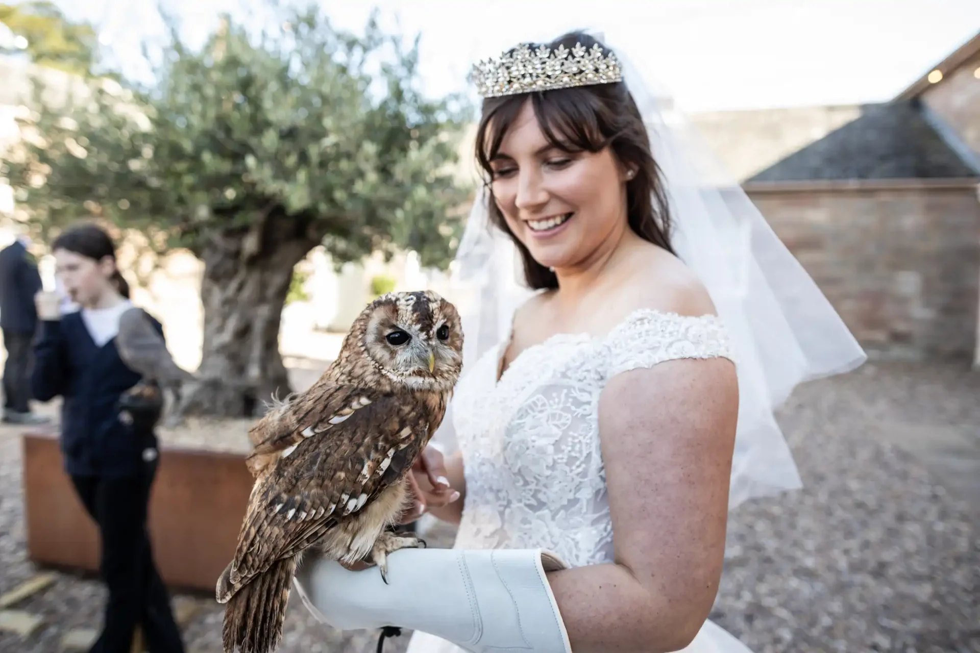 Bride in a white dress and tiara holds a brown owl on her gloved hand outdoors.