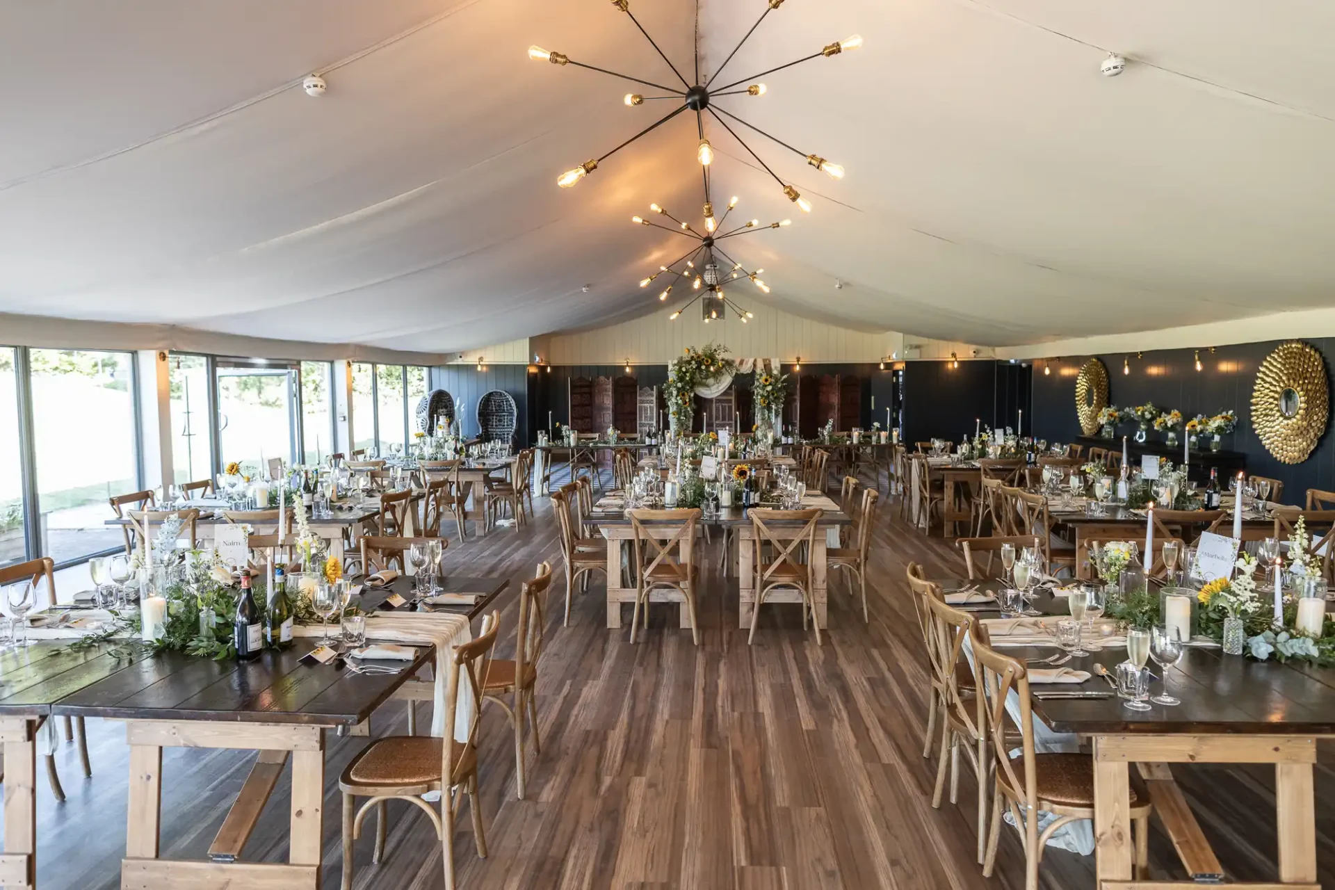 Spacious event hall with wooden flooring, set with rows of wooden tables and chairs. Chandeliers hang from the ceiling, and floral decorations adorn the tables. Large windows are on one side.