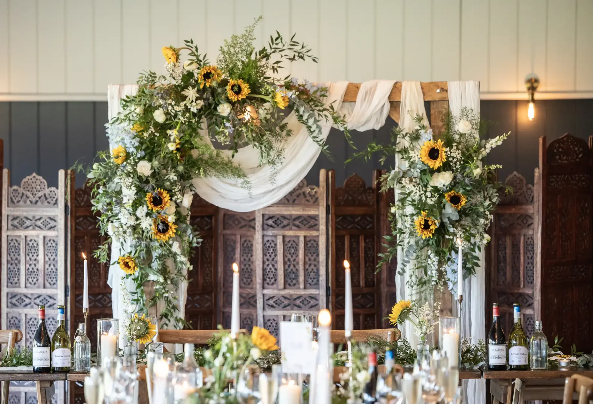 Floral wedding arch with sunflowers and white drapery, set behind a decorated table with candles and wine bottles.