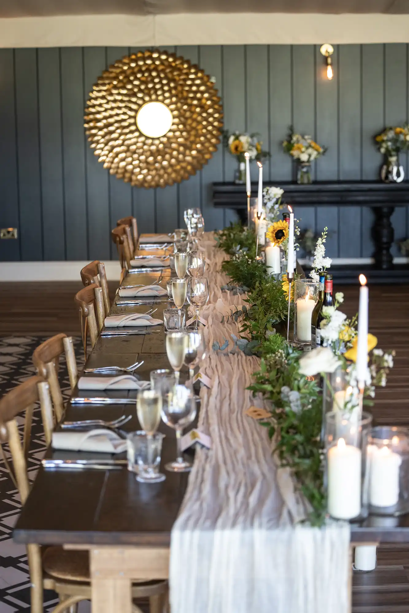 Elegant dining table setting with a long, draped runner, greenery, candles, and sunflowers. Chairs line both sides, and champagne glasses are arranged. A large mirror decorates the wall.