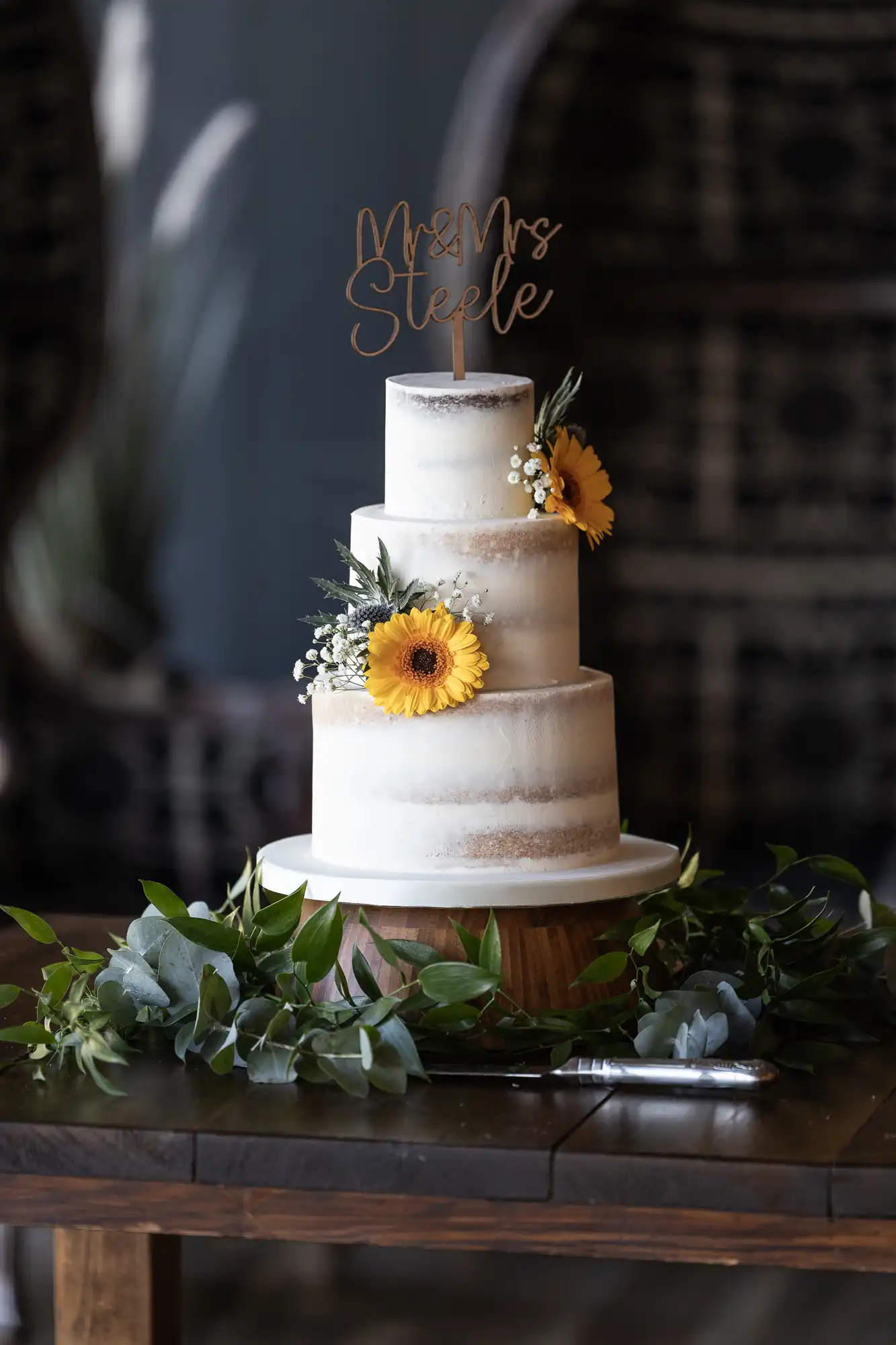 Three-tier semi-naked cake with "Mr & Mrs Steele" topper, decorated with sunflowers and greenery, on a wooden table with foliage.