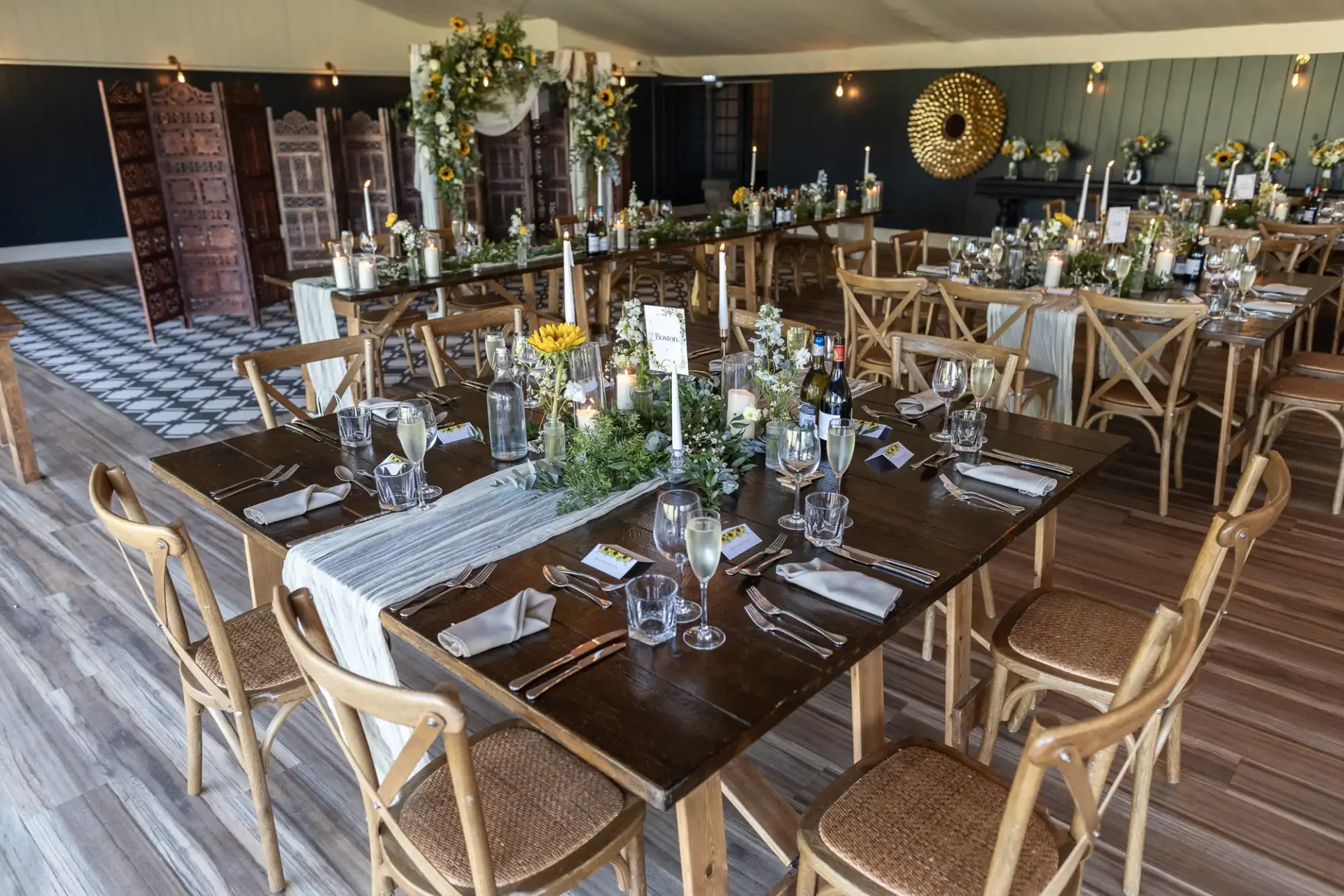 Rustic wedding reception setup with long wooden tables, elegant table settings, sunflowers, and greenery. Wooden chairs surround tables under warm ambient lighting.