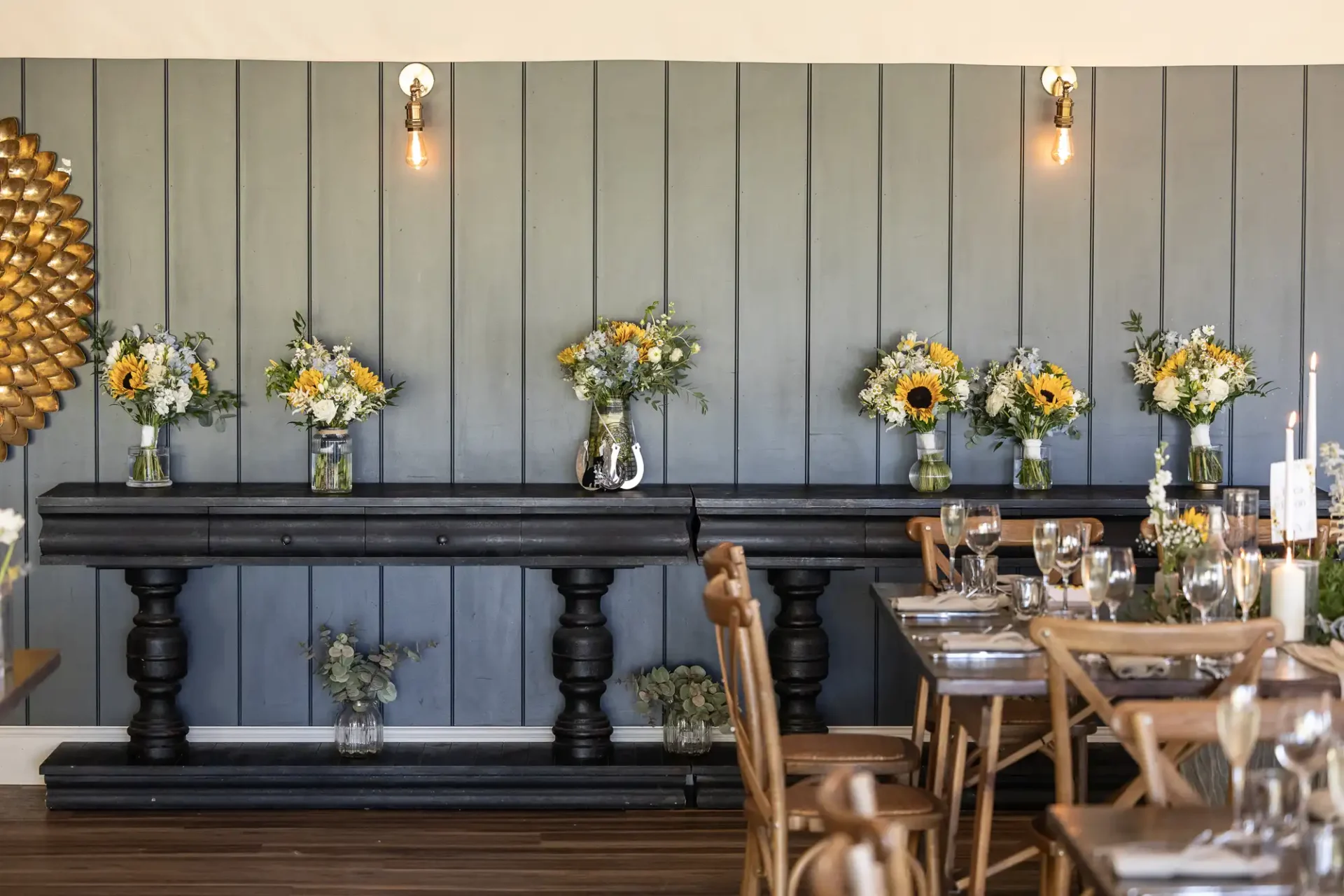 A rustic dining area features a wooden table with neatly arranged place settings. Sunflower bouquets in glass vases adorn a dark side table against a gray paneled wall with overhead lights.