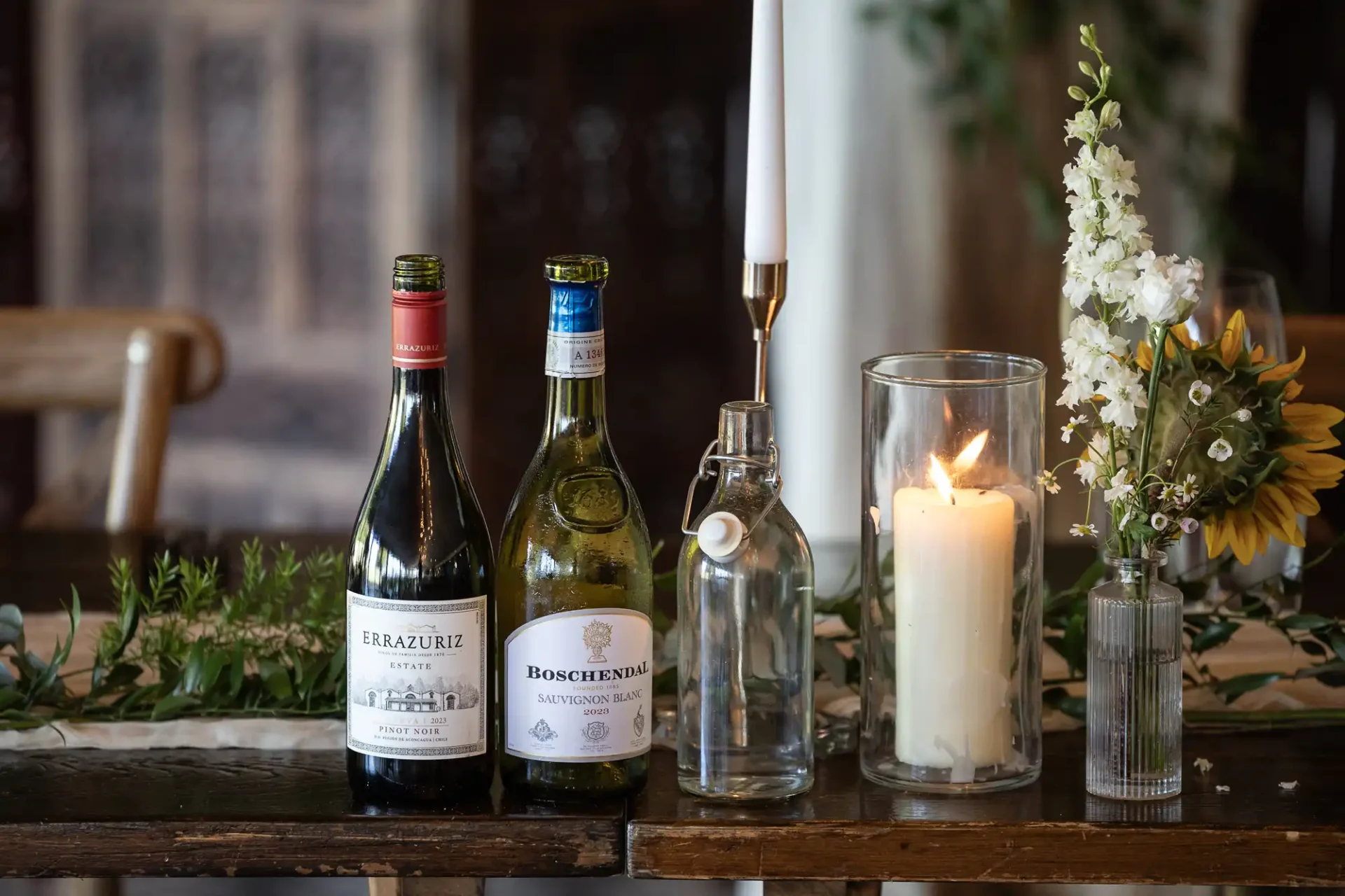 Two wine bottles, a candle in a glass holder, an empty bottle, and flowers are arranged on a wooden table with greenery.