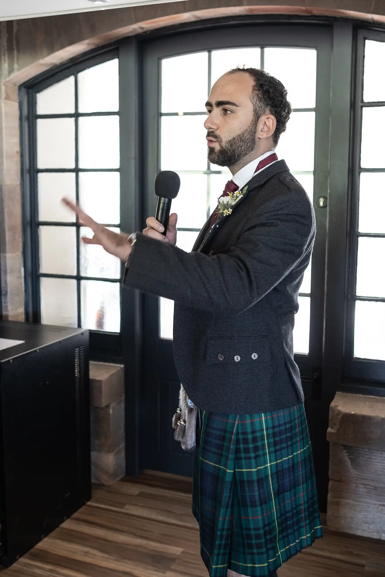 Man in a kilt holding a microphone, speaking indoors in front of large windows.