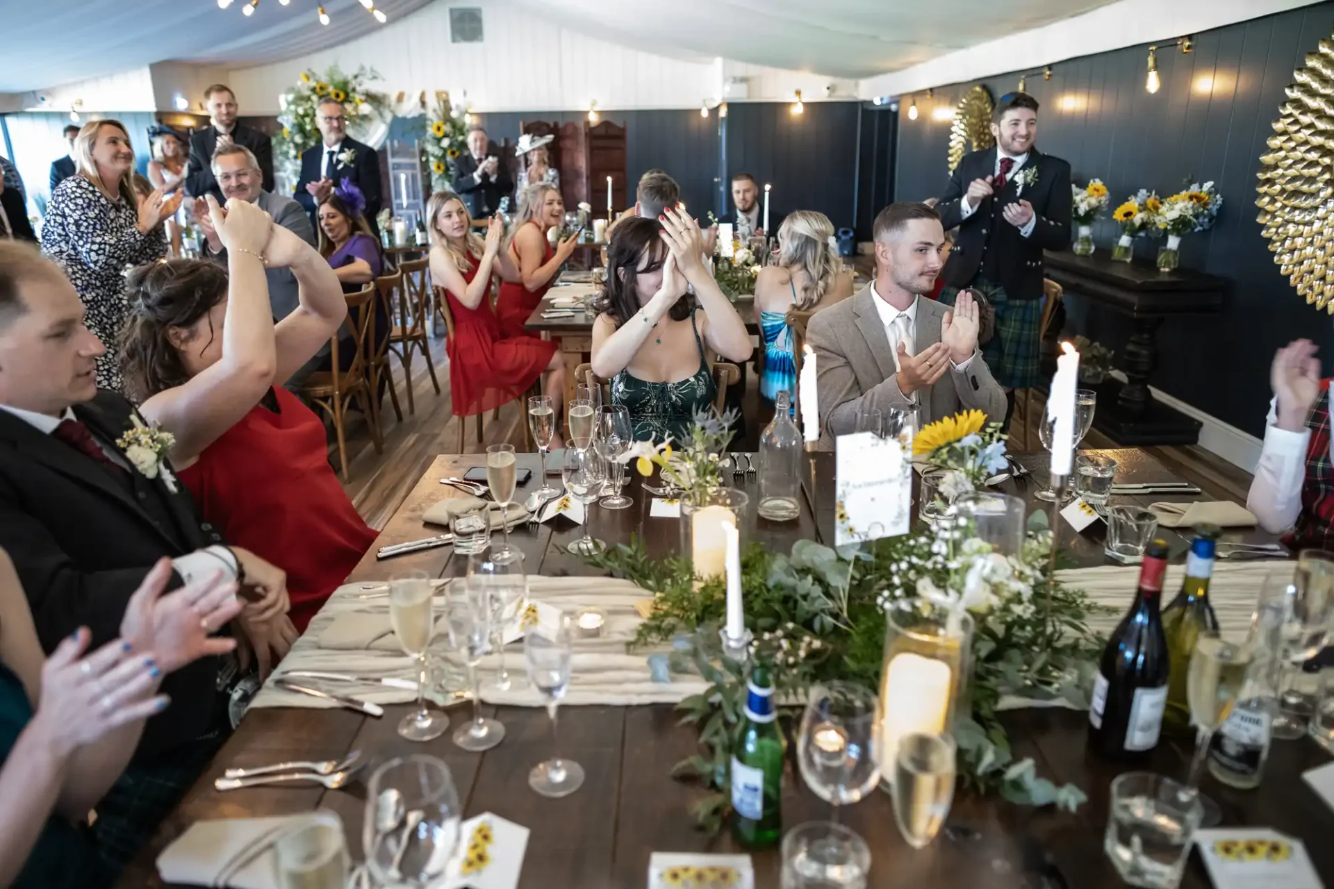 Guests clapping at a formal event in a decorated venue with a long dining table, candles, flowers, and drinks.