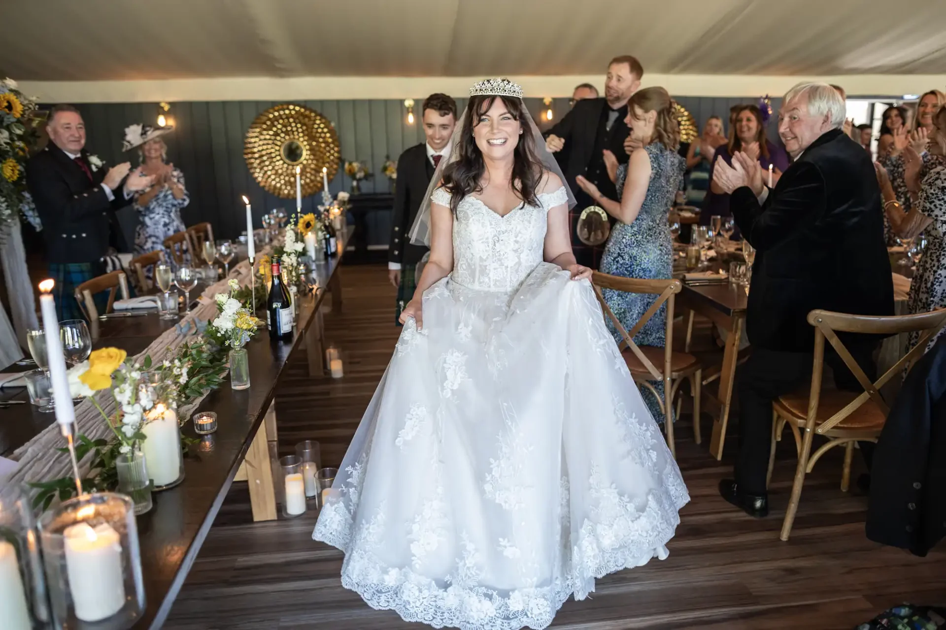 A bride, wearing a crown and white gown, walks between tables in a decorated venue. Guests are clapping and smiling around her.