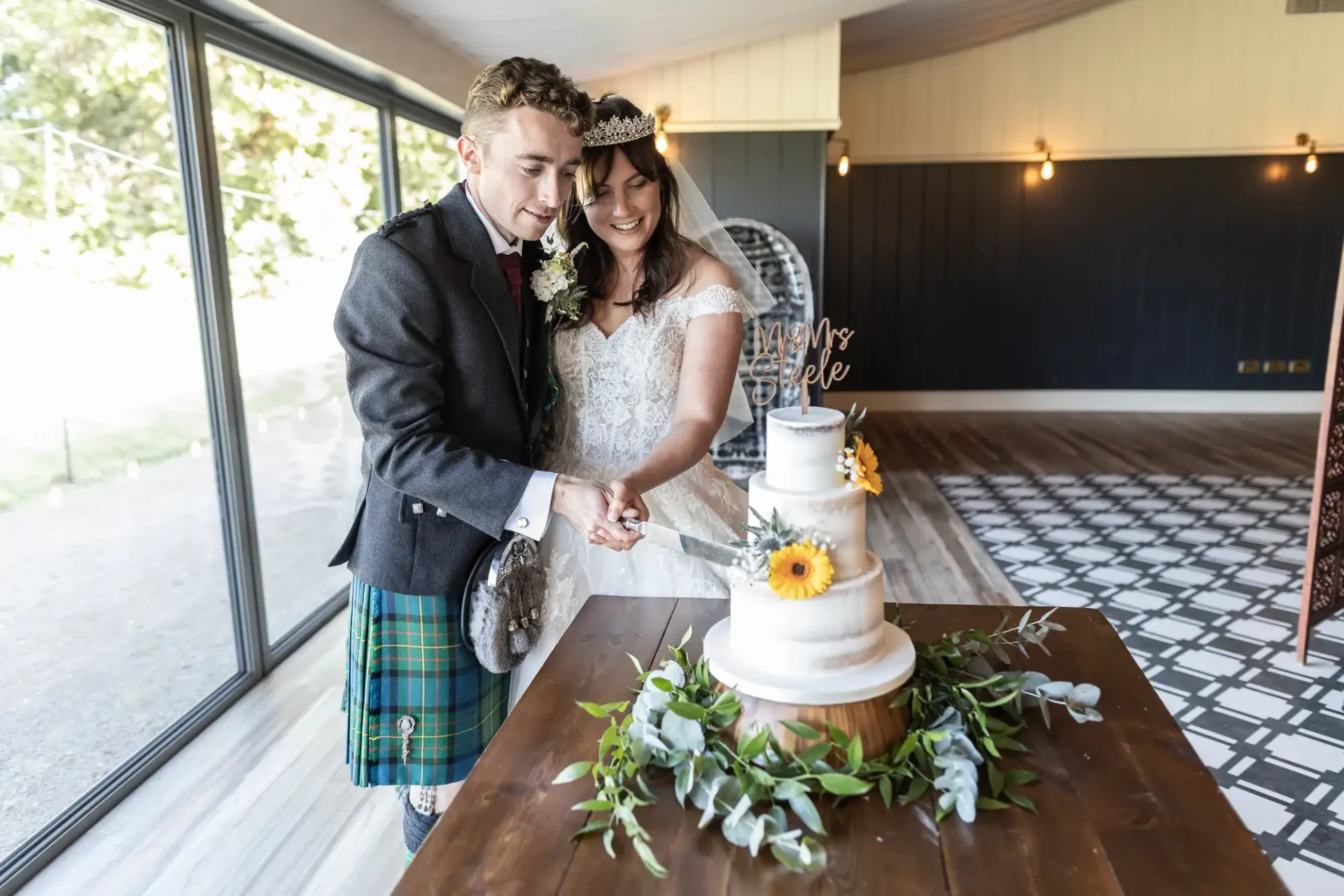 A bride and groom cut a three-tiered wedding cake decorated with flowers. The groom wears a kilt, and the bride is in a white dress and tiara. They are in a room with large windows.