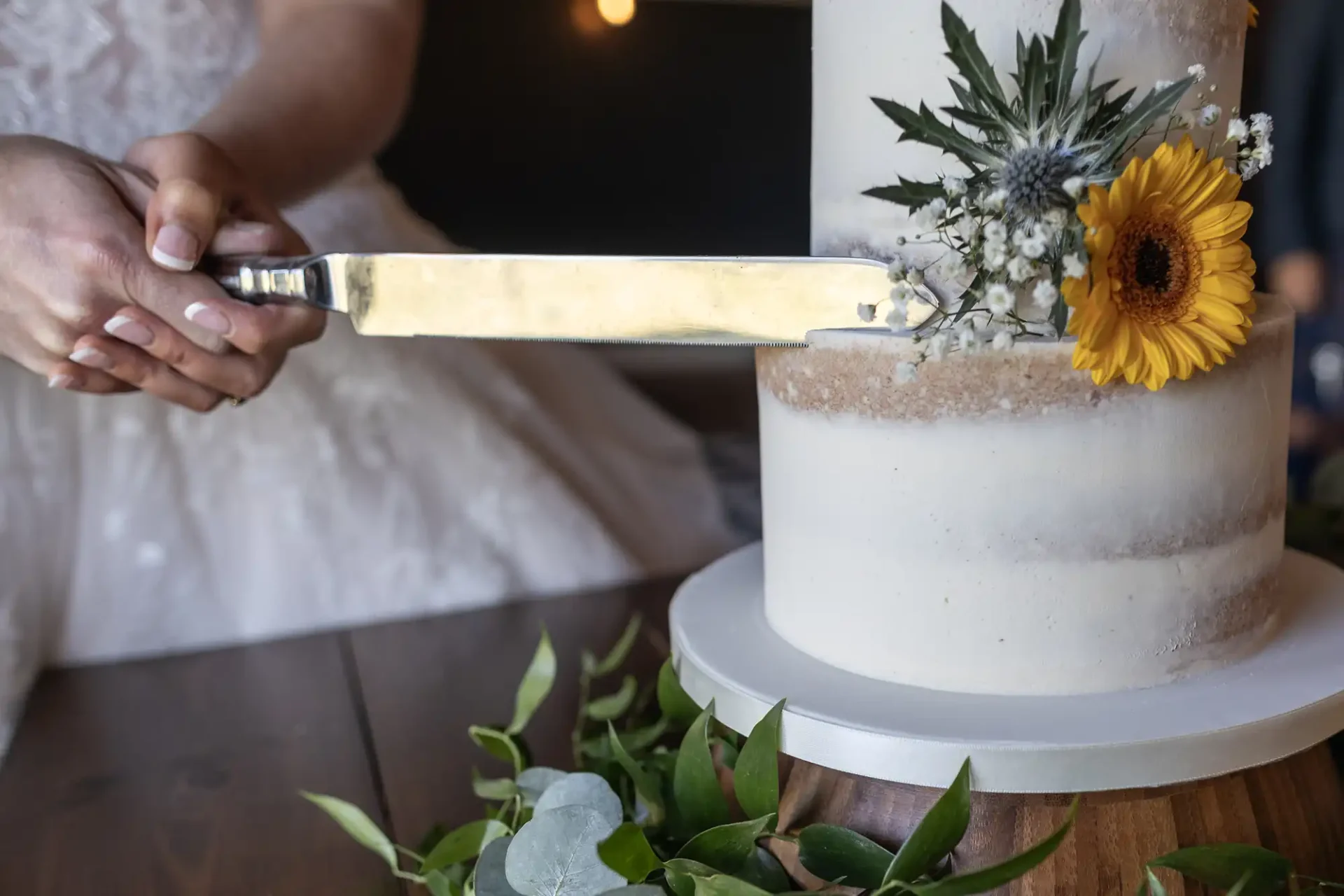 Person cutting a white-tiered cake adorned with a sunflower and greenery, using a large knife.