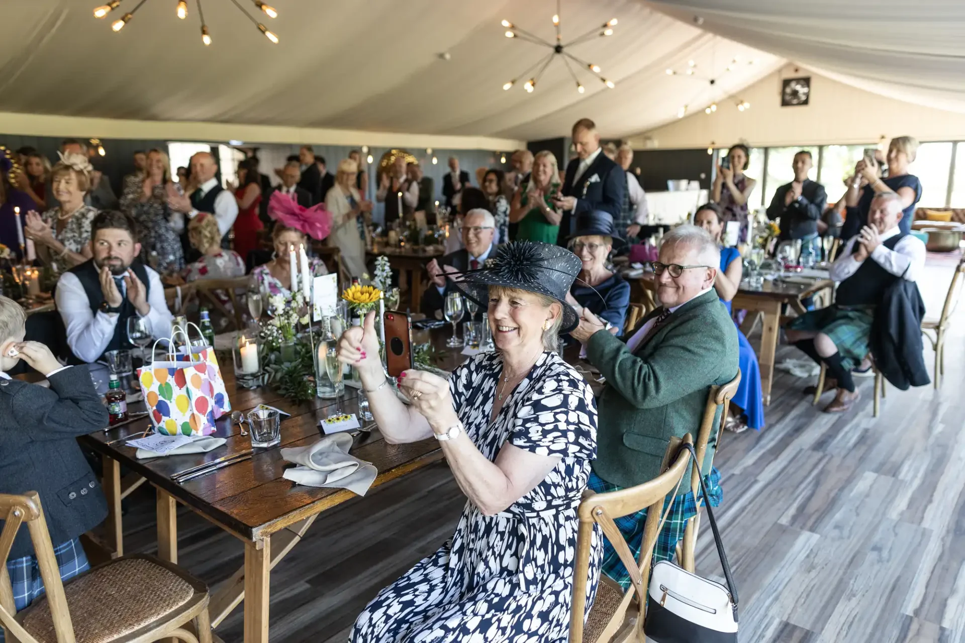 Guests seated and standing in a decorated venue, many clapping and taking photos. A woman in a patterned dress and hat holds a phone, capturing the moment.