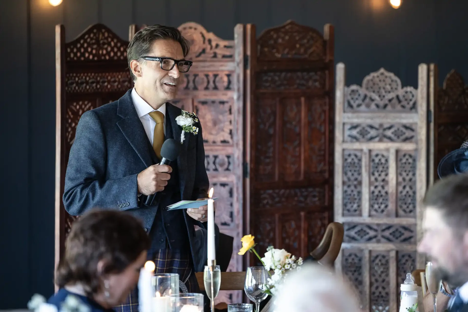 Man in a suit holding a microphone and notecards, smiling while speaking at an event. Decorative wooden screens are in the background.