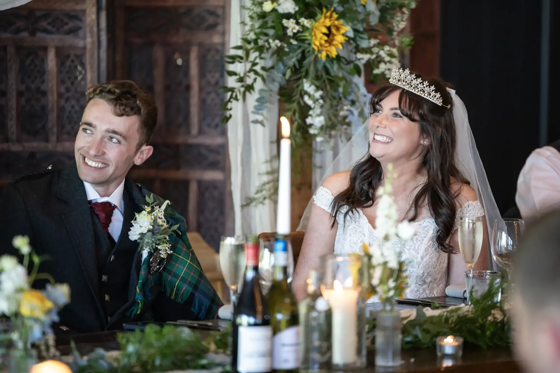 Bride and groom smiling at a decorated table, featuring candles, flowers, and greenery. Bride wears a tiara and veil; groom wears a suit with a tartan sash.