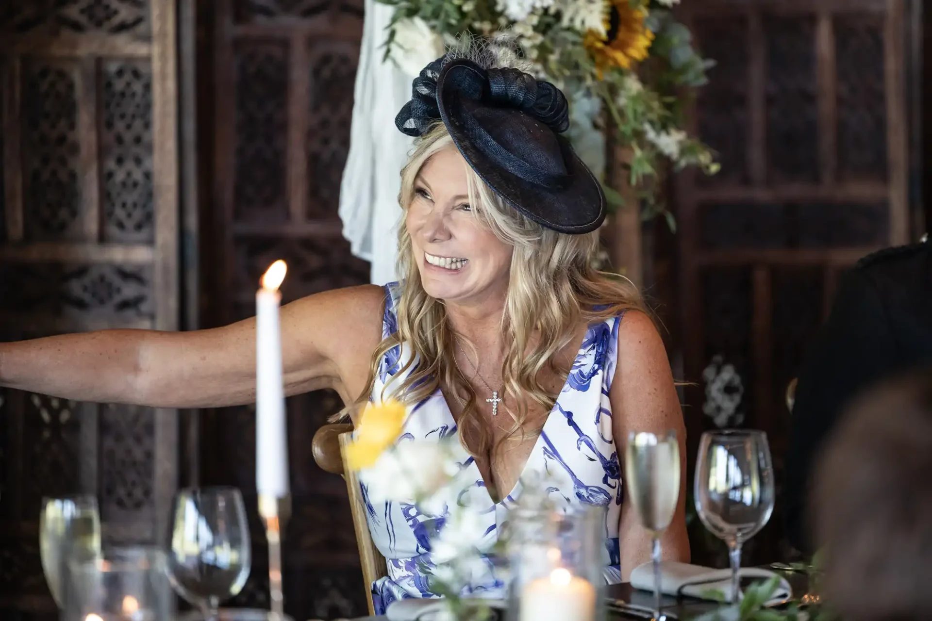 Woman in a floral dress and hat smiles and gestures while seated at a decorated table with candles and flowers.