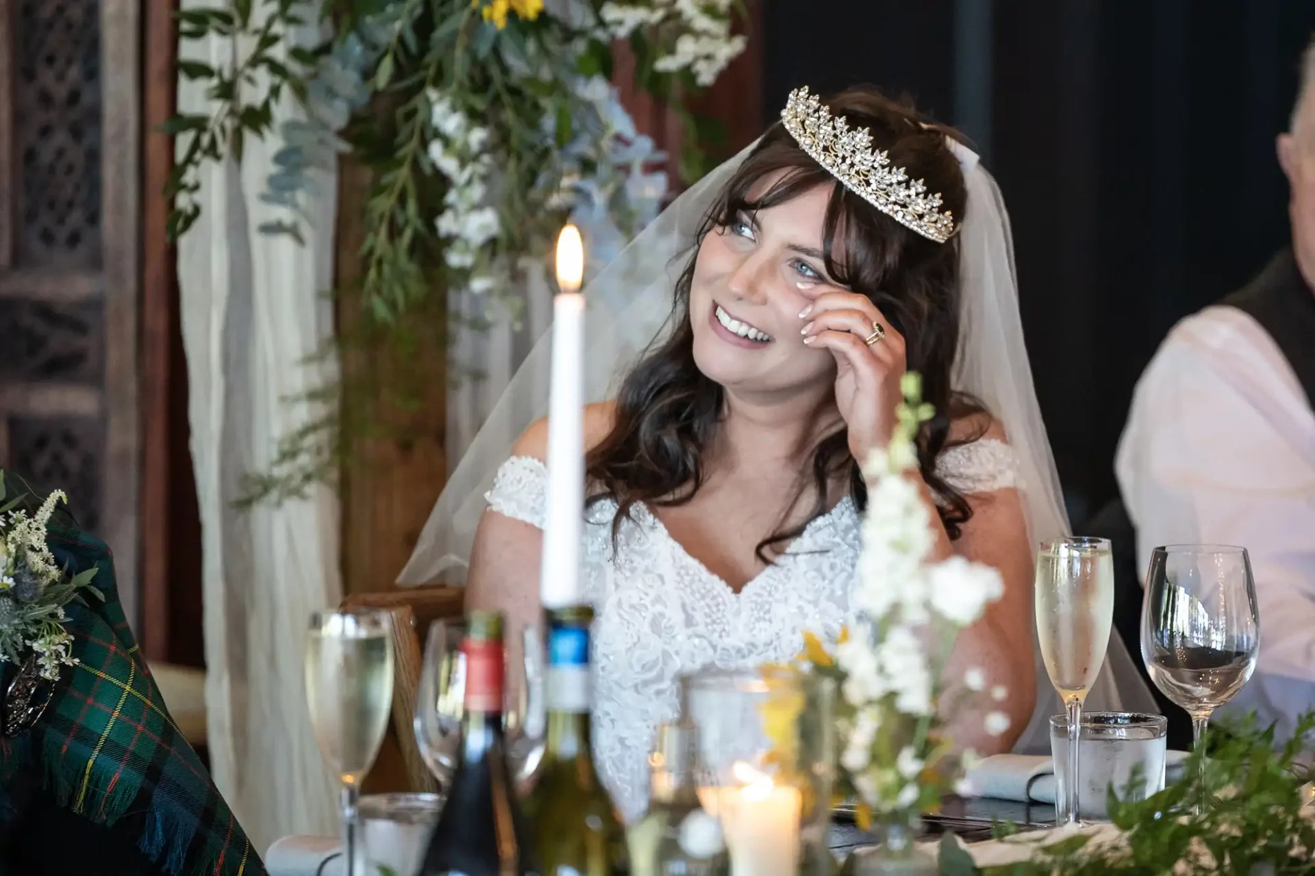 Bride in a white dress and tiara smiles while seated at a table with floral decorations, candles, and wine glasses.