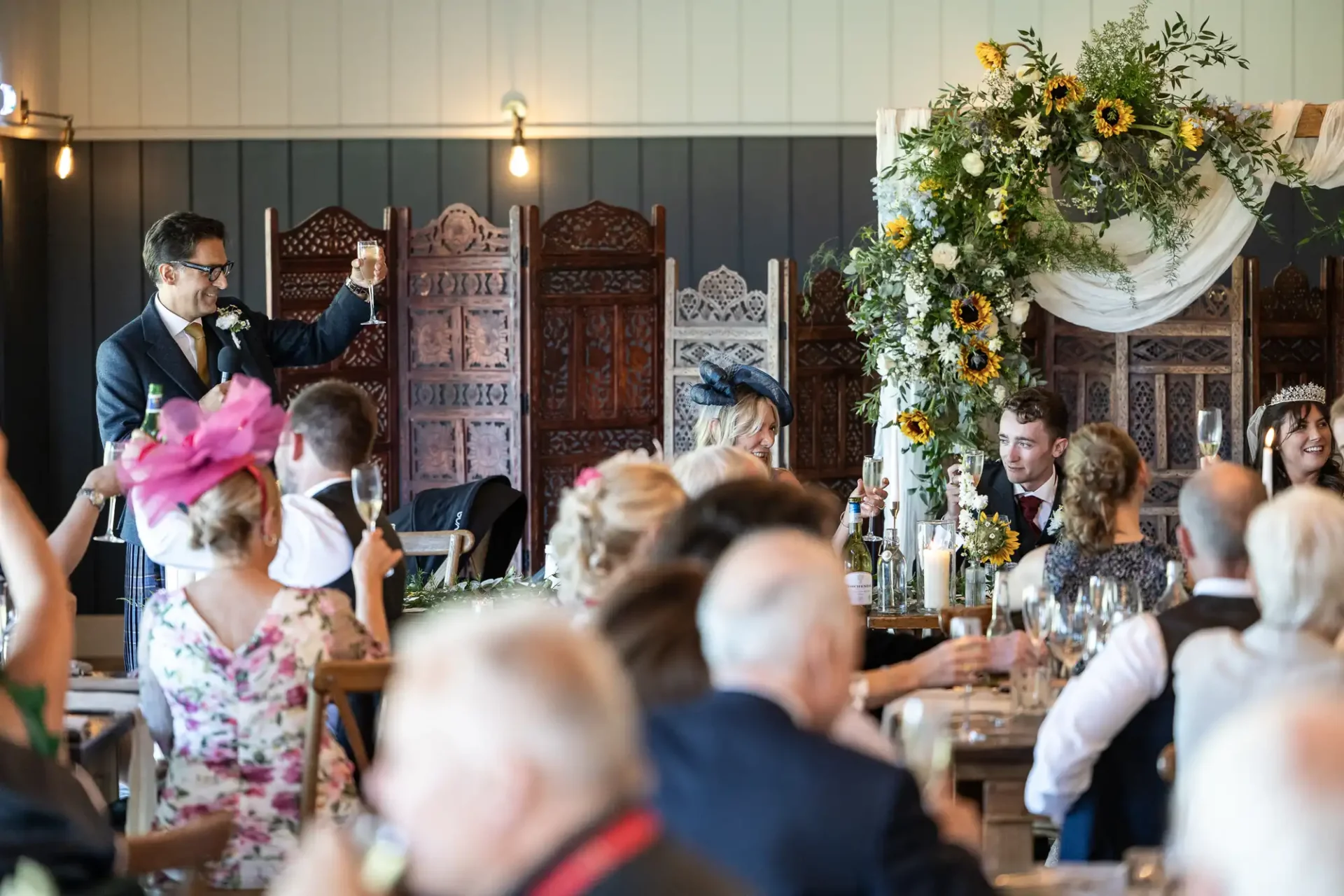 A man gives a toast at a wedding reception, while guests sit at decorated tables. Sunflowers and greenery adorn an arch behind the couple.