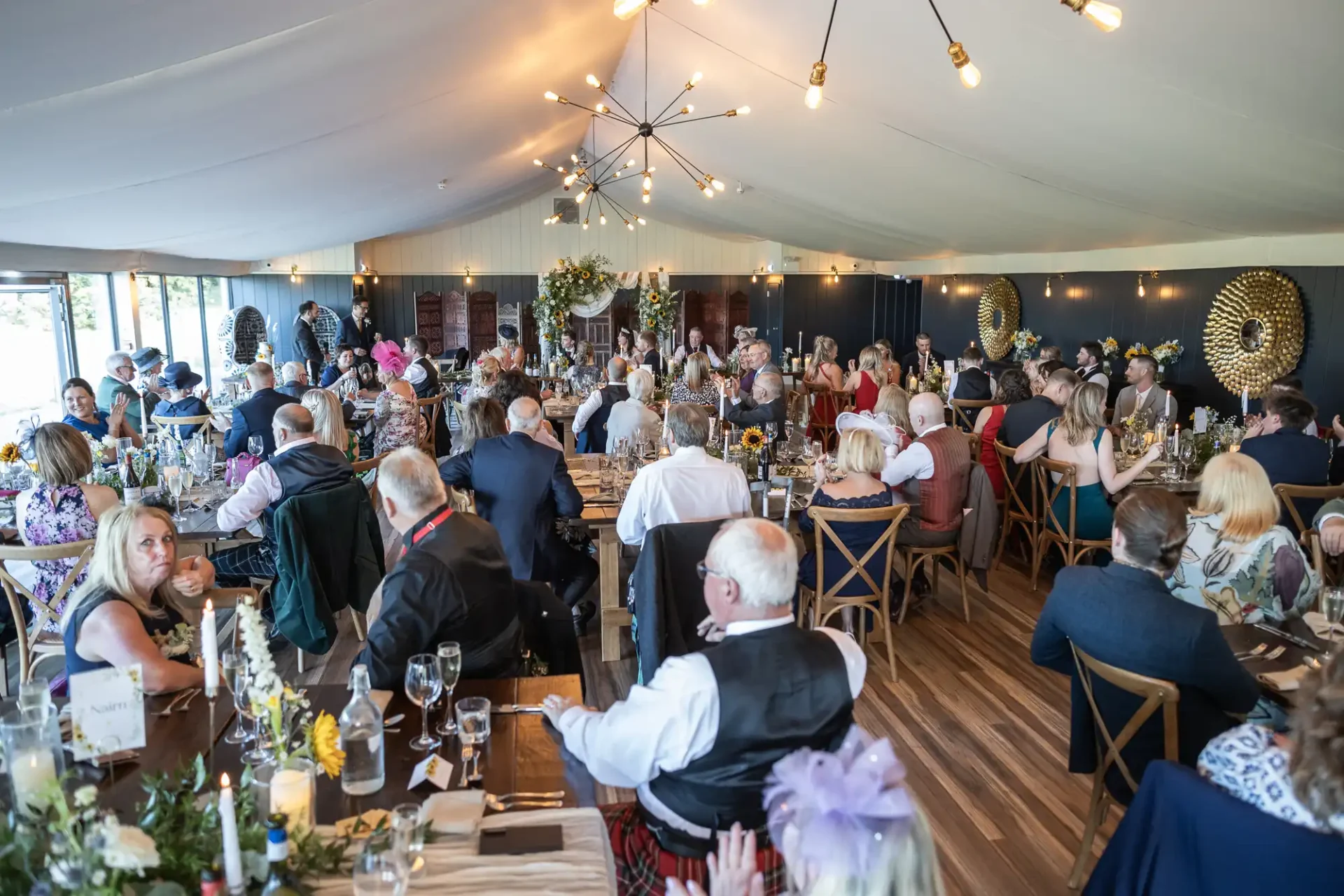 A large group of people seated at tables inside a decorated tent, likely attending a formal event or celebration. The room is well-lit with chandeliers and floral arrangements.