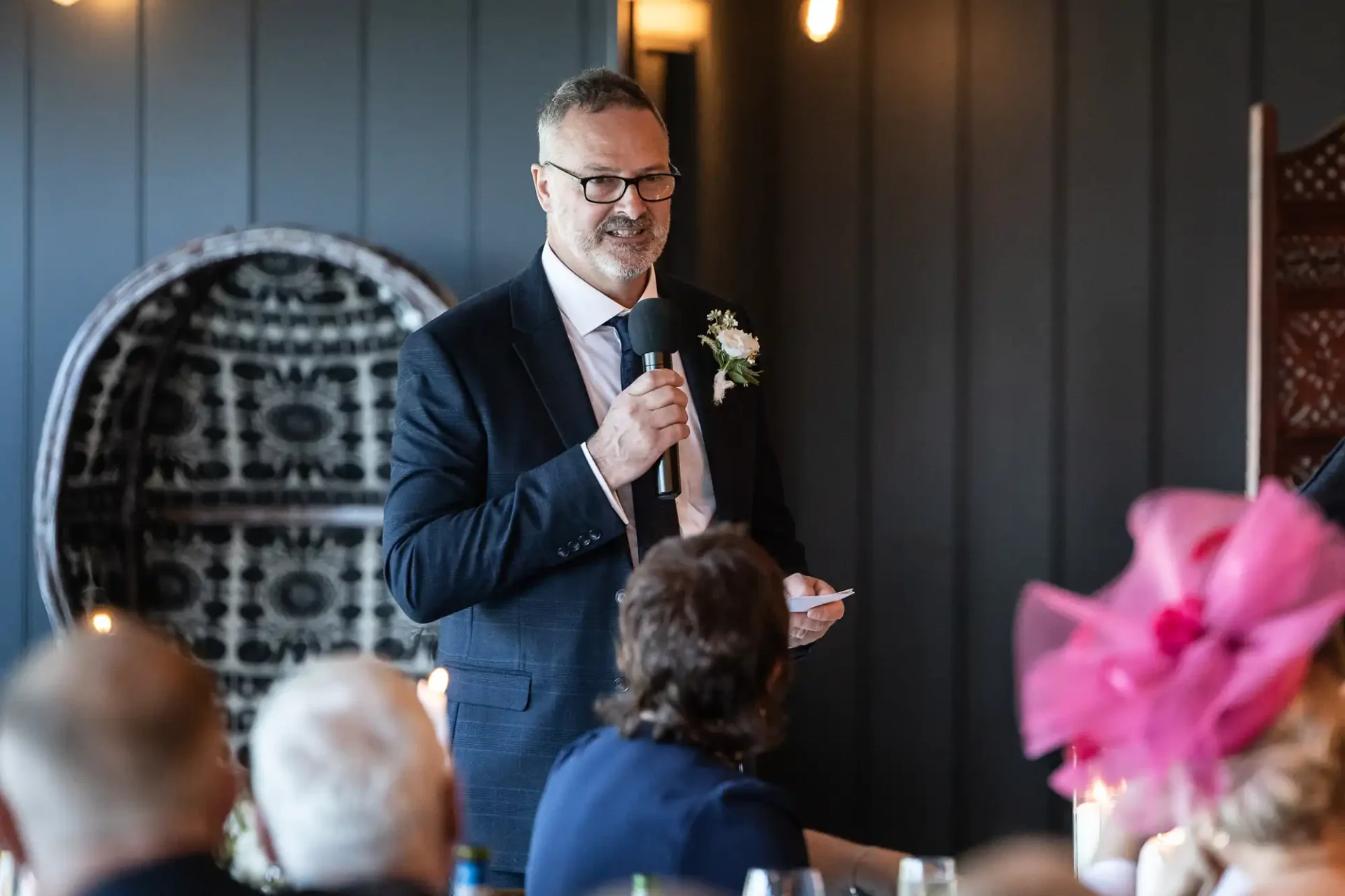 A man in a suit and glasses is giving a speech with a microphone in hand. He has a flower on his lapel. People are seated in front of him.