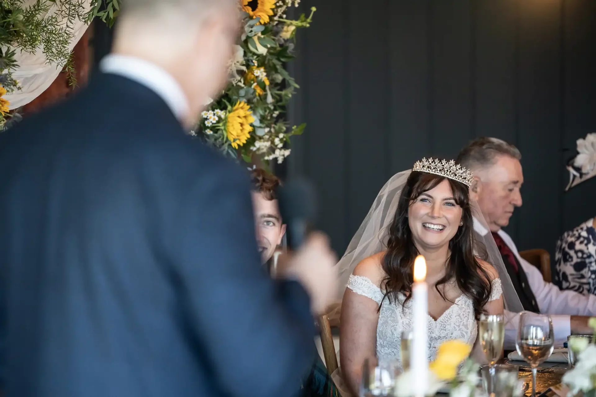 A bride wearing a tiara and veil smiles at a man speaking into a microphone during a wedding reception.